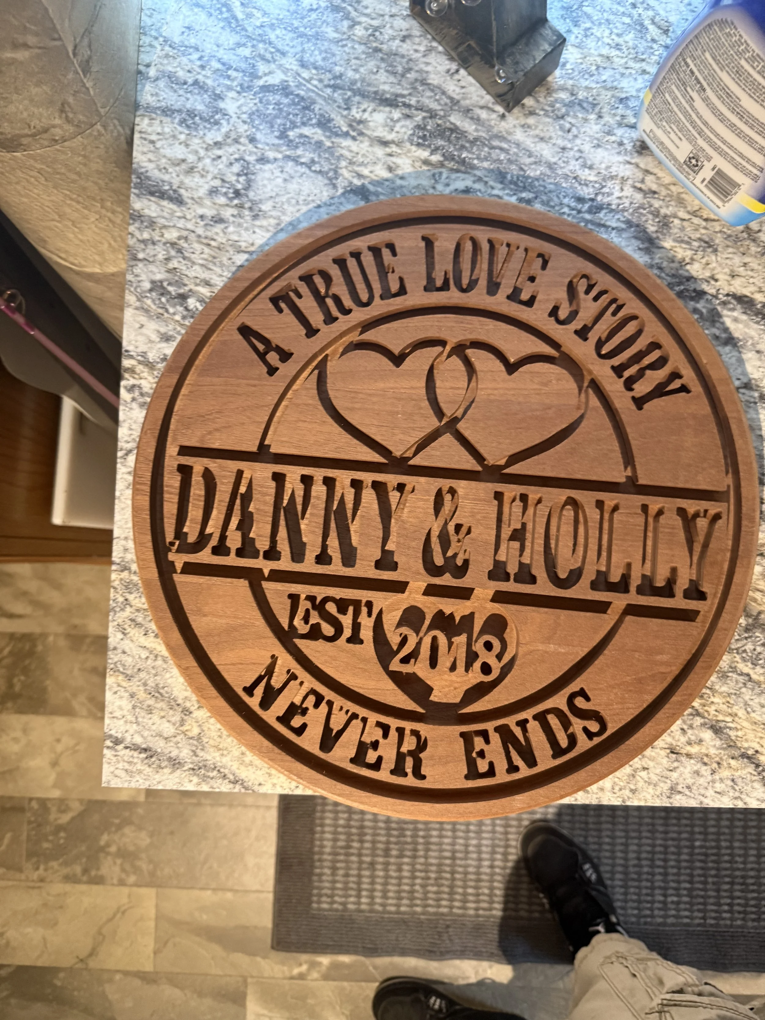 Wooden decorative plaque with engraved text: "A TRUE LOVE STORY DANNY & HOLLY EST. 2018 NEVER ENDS" on a granite countertop.