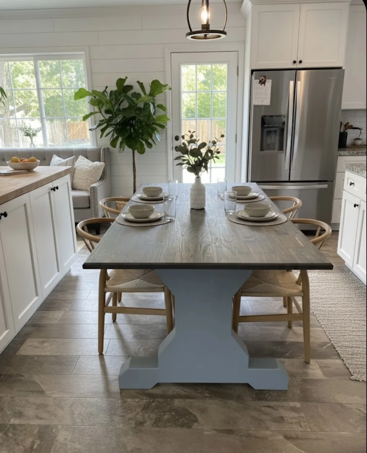 A dining area inside a bright, modern kitchen with a wooden table set with four place settings, a white vases with greenery, a large indoor plant, white kitchen cabinets, and a stainless steel refrigerator. Natural light is coming through windows and