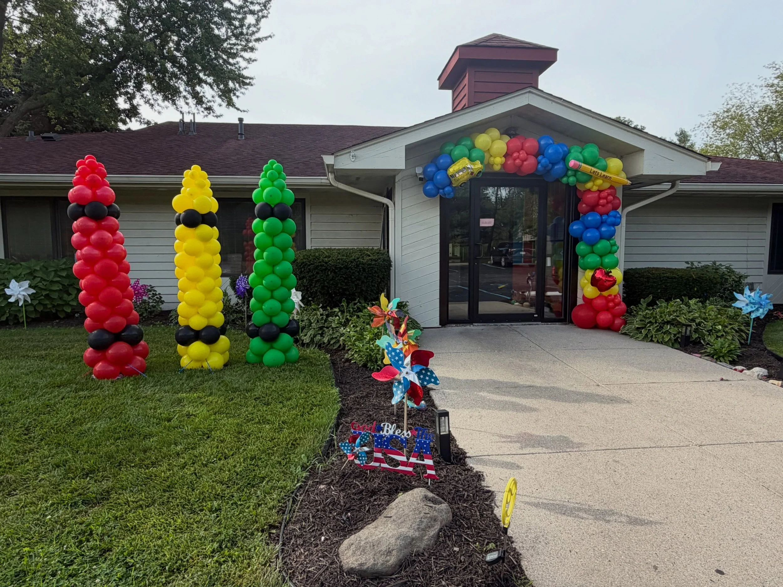 Building entrance decorated with colorful balloon columns, a balloon arch, and patriotic garden decorations