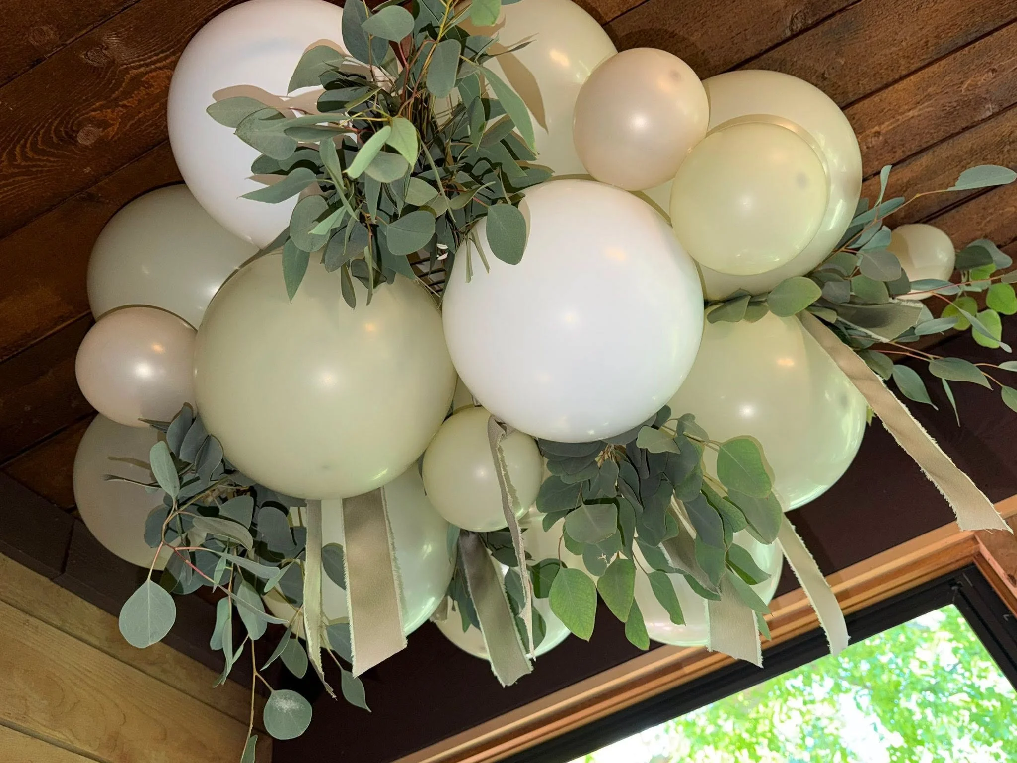 Hanging arrangement of white balloons, eucalyptus greenery, and flowing cream ribbons beneath a wooden ceiling