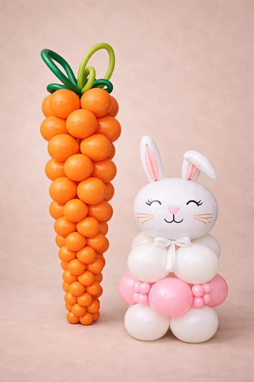 Orange balloon carrot with green leaves beside a white and pink bunny balloon on a pastel pink background
