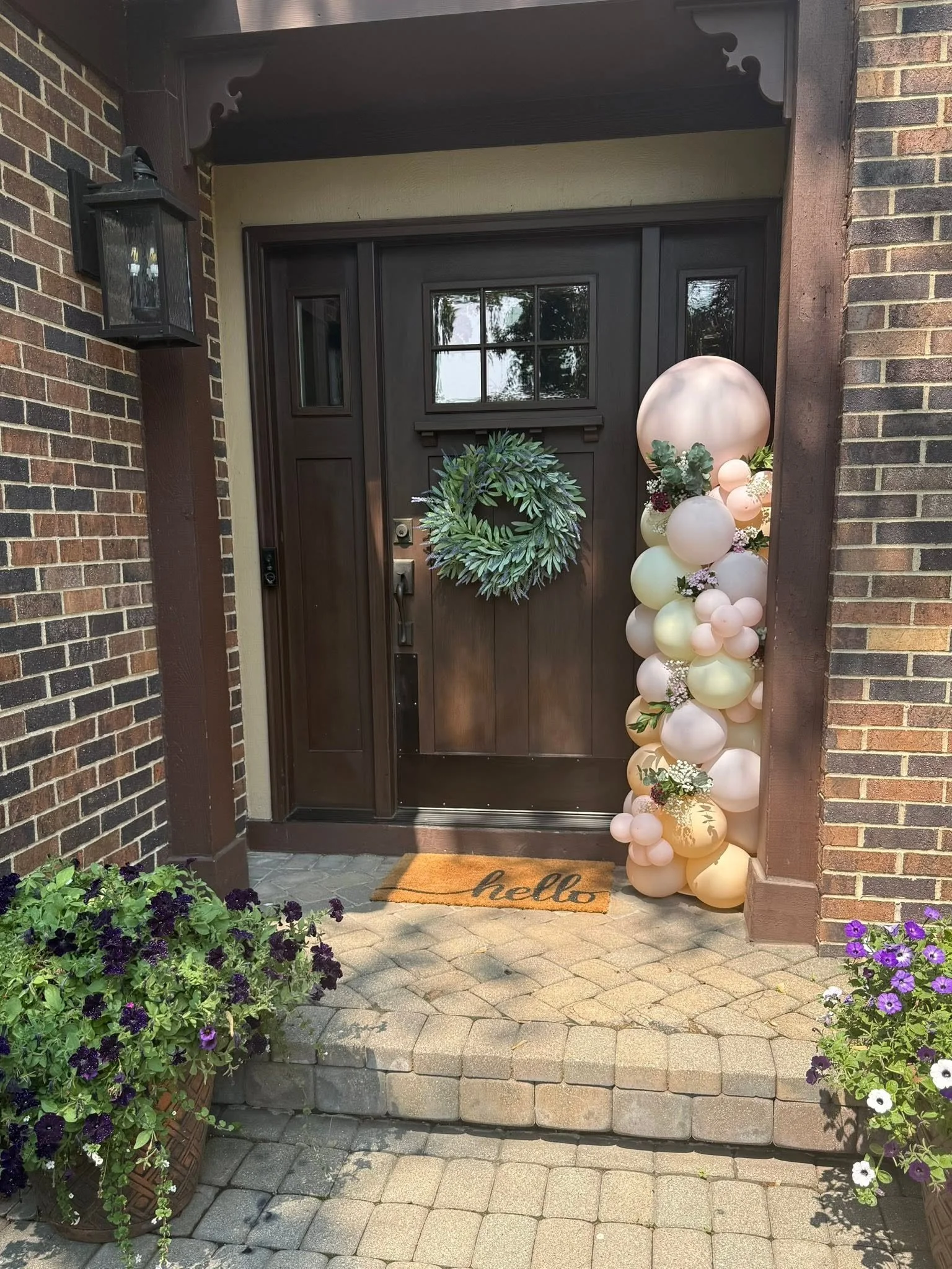 Dark wood front door with green wreath, pastel balloon garland, floral planters, and a welcome mat.