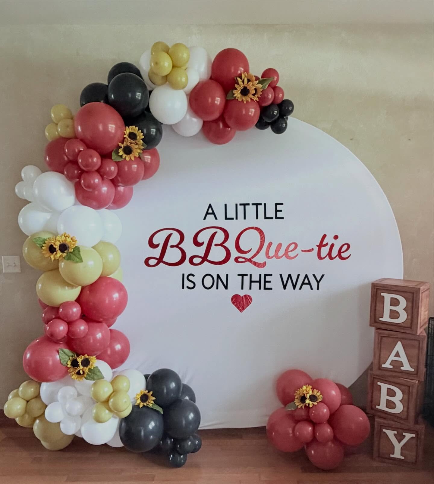 Baby shower backdrop with a colorful balloon arch, sunflowers, letter blocks, and sign reading “A Little BBQ-ue-tie Is on the Way.”