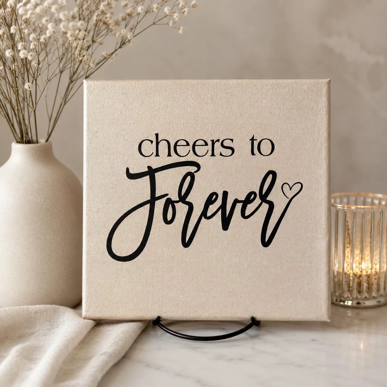 Neutral wedding decor featuring a canvas sign reading “Cheers to Forever” with a heart, beside a vase of dried flowers and a candle lantern.