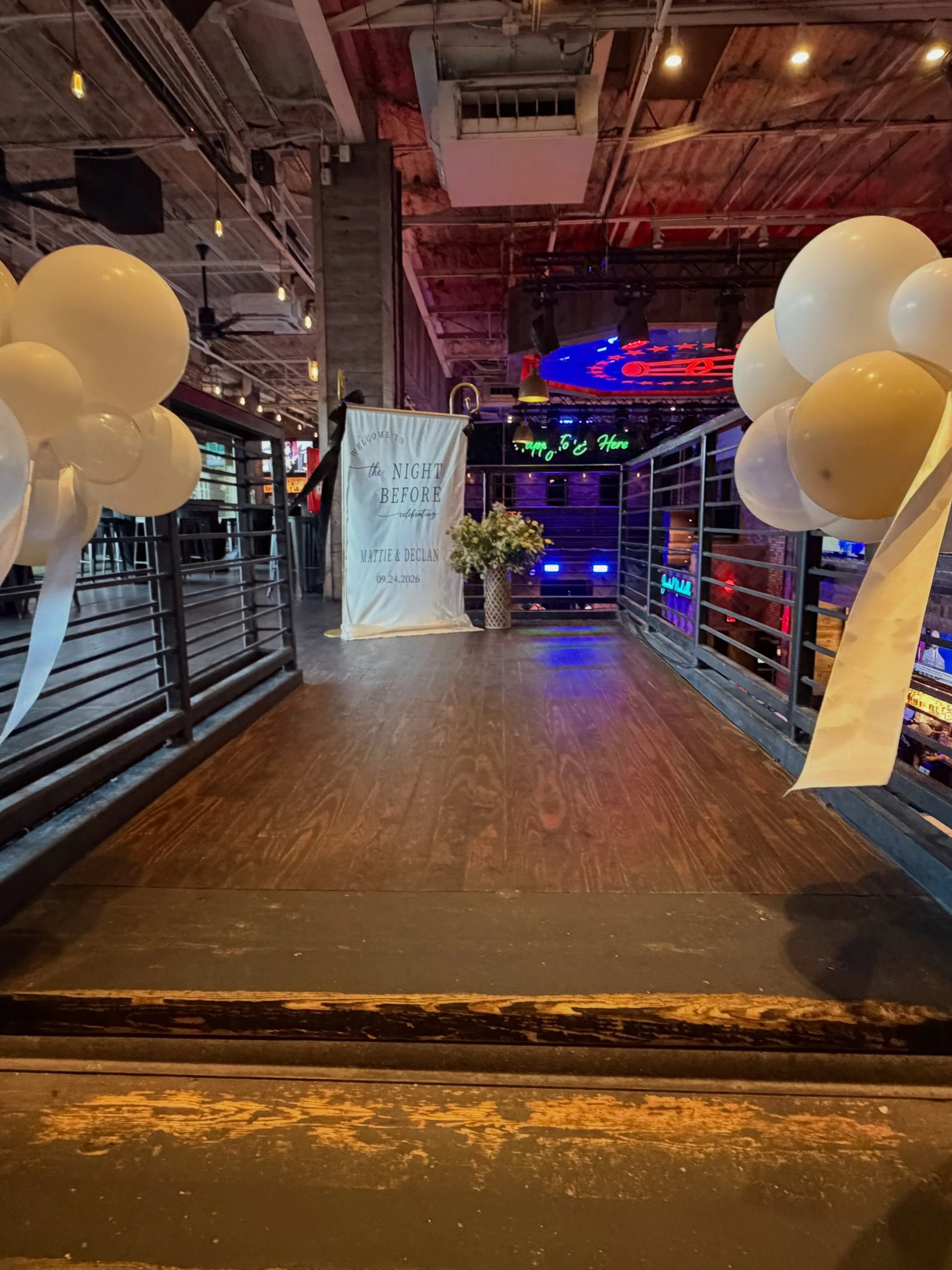 Decorated event venue with cream balloons, a welcome-to-the-night-before wedding banner, DJ stage lighting, and industrial loft ceiling