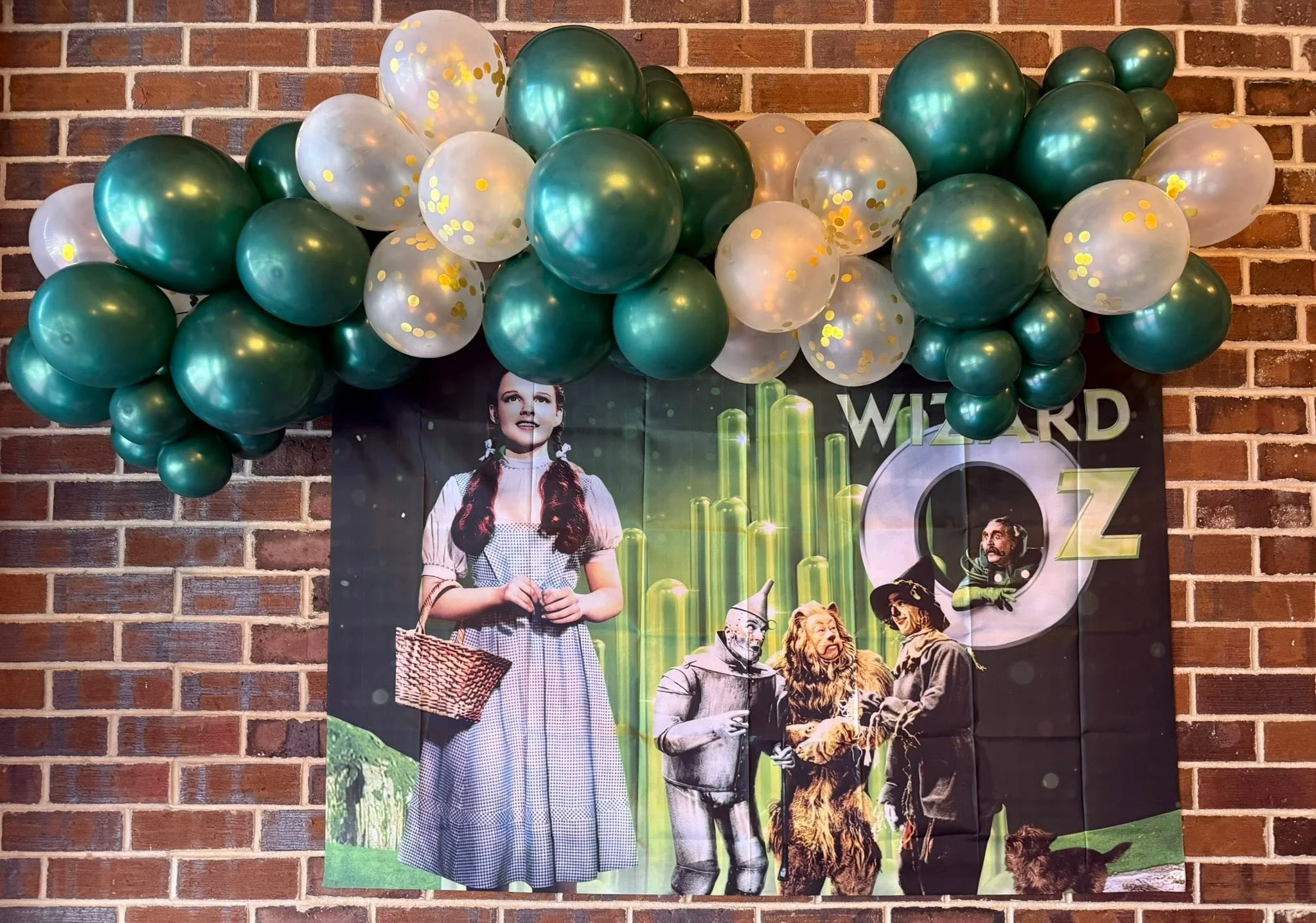 Teal and white balloon garland with gold confetti decorating a Wizard of Oz poster on a brick wall