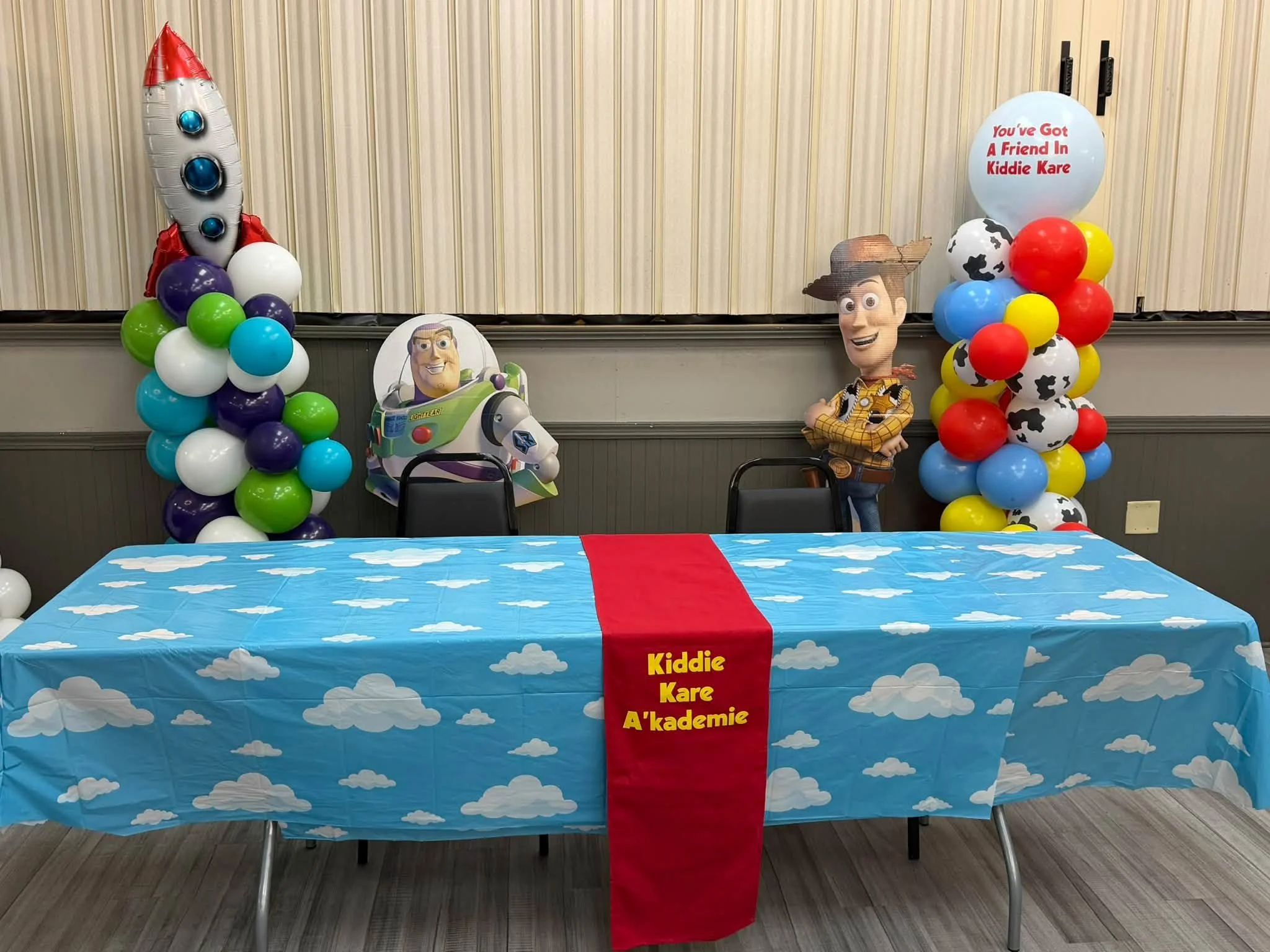 Toy Story–themed party table with Buzz Lightyear and Woody decorations, colorful balloon columns, and a cloud-print tablecloth labeled Kiddie Kare A’kademie