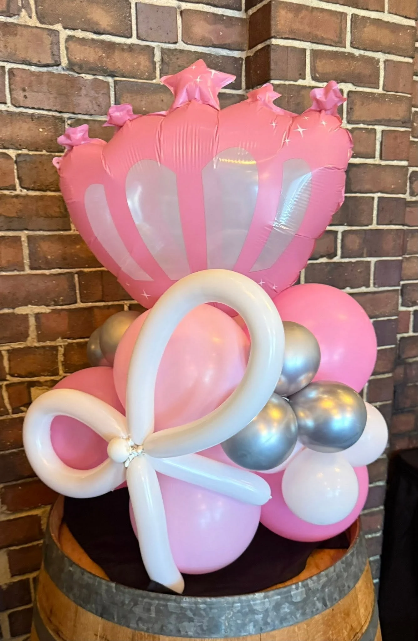 Pink, white, and silver balloon arrangement with a large crown-shaped balloon, displayed on a wooden barrel against a brick wall.