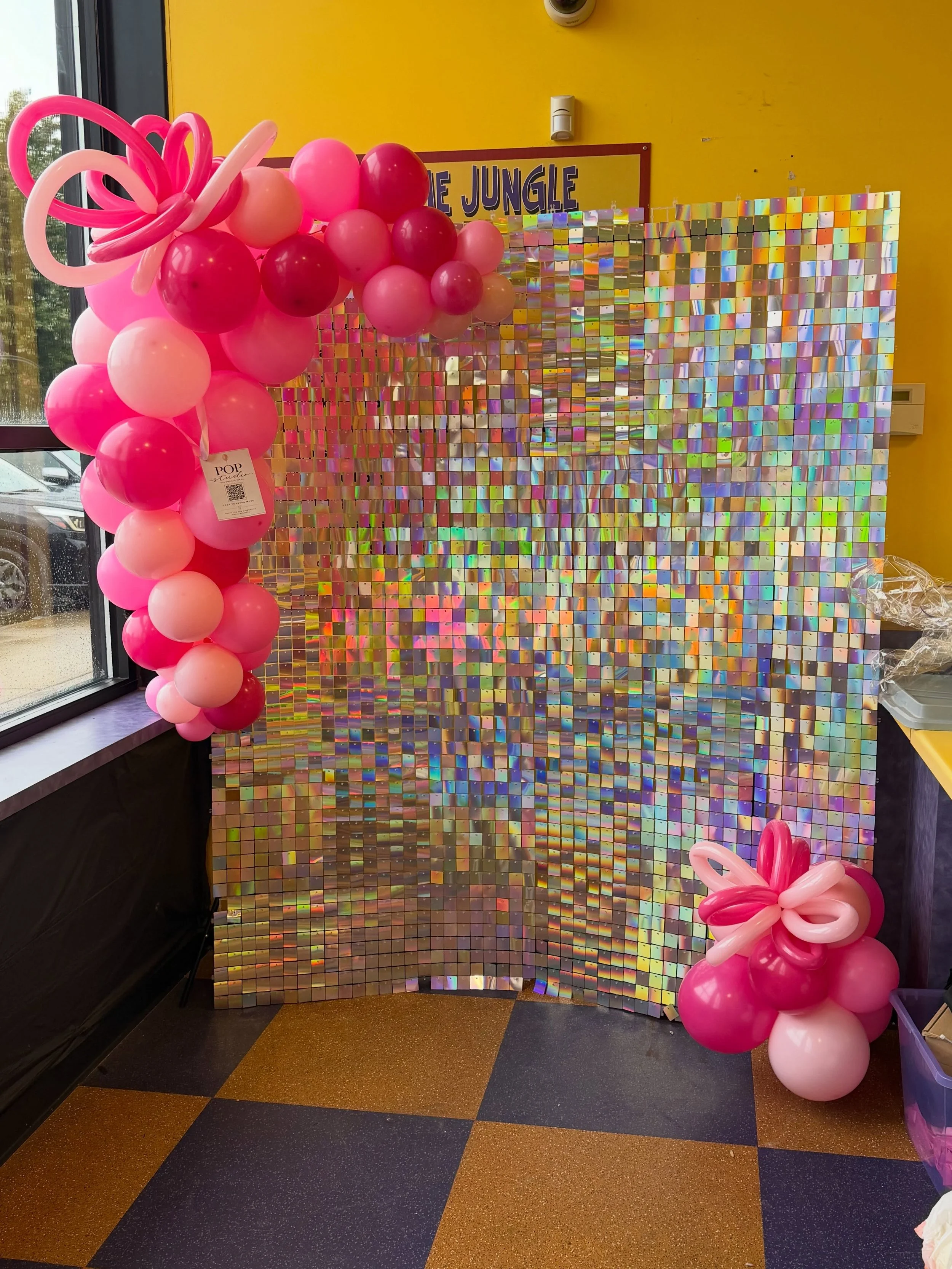 Pink balloon clusters with twisted balloon flowers beside a shimmering iridescent sequin photo backdrop in a colorful event space.