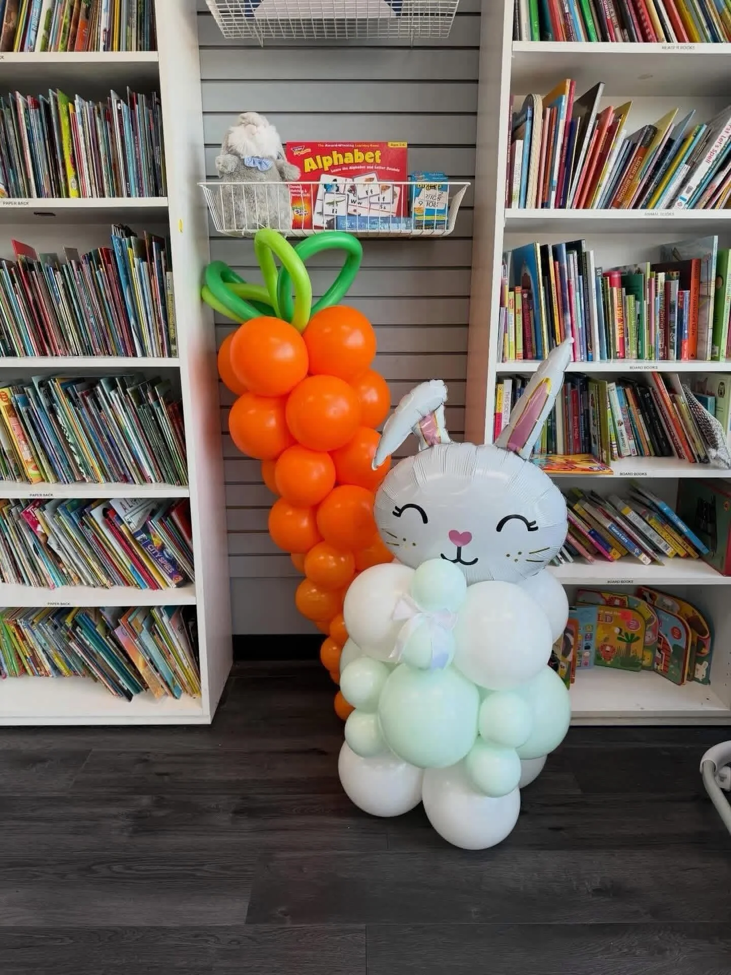 Pastel balloon cat and orange balloon bouquet displayed in a children's bookstore surrounded by bookshelves
