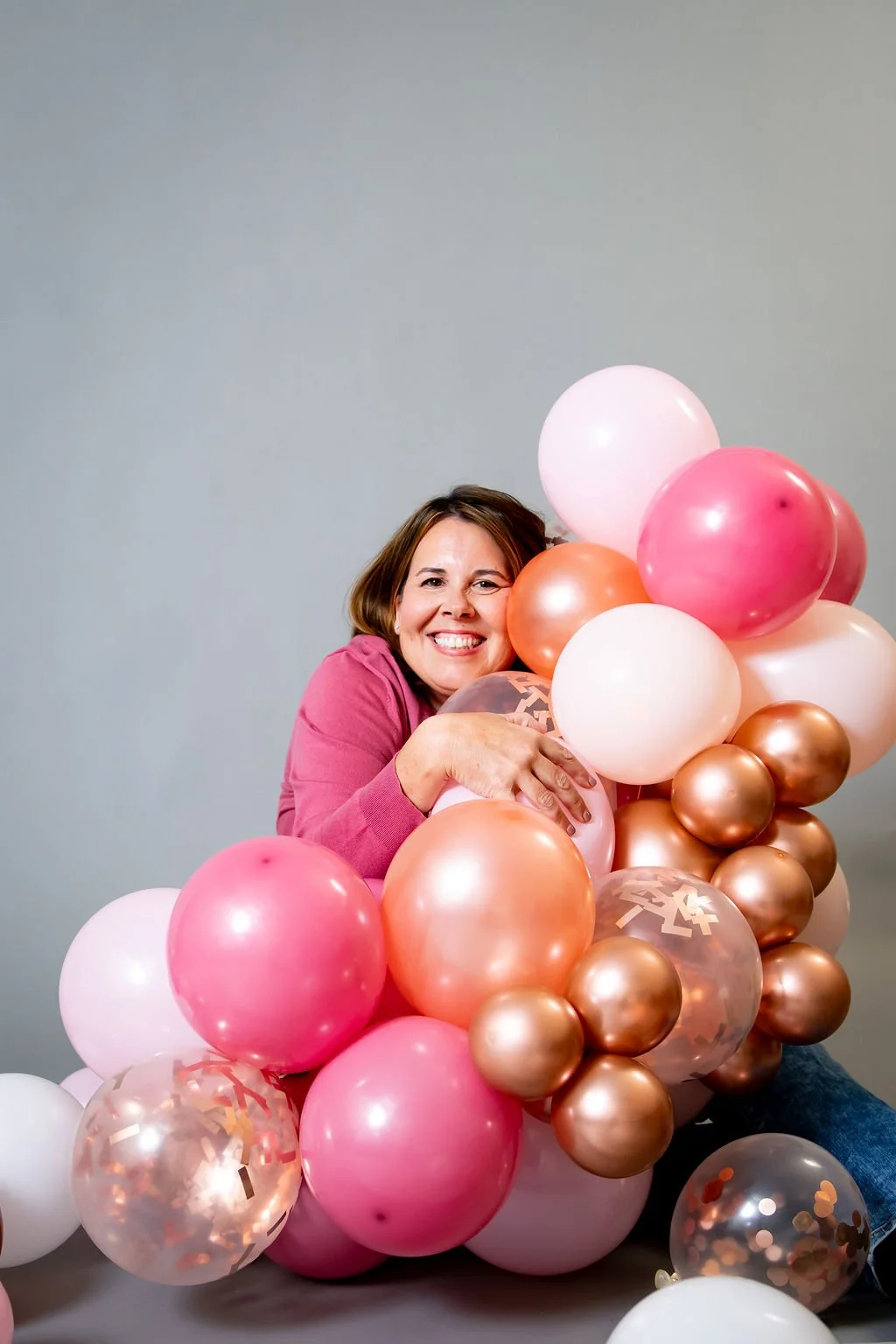 Smiling woman in a pink sweater holding a colorful pink, peach, white, and rose-gold balloon arrangement.
