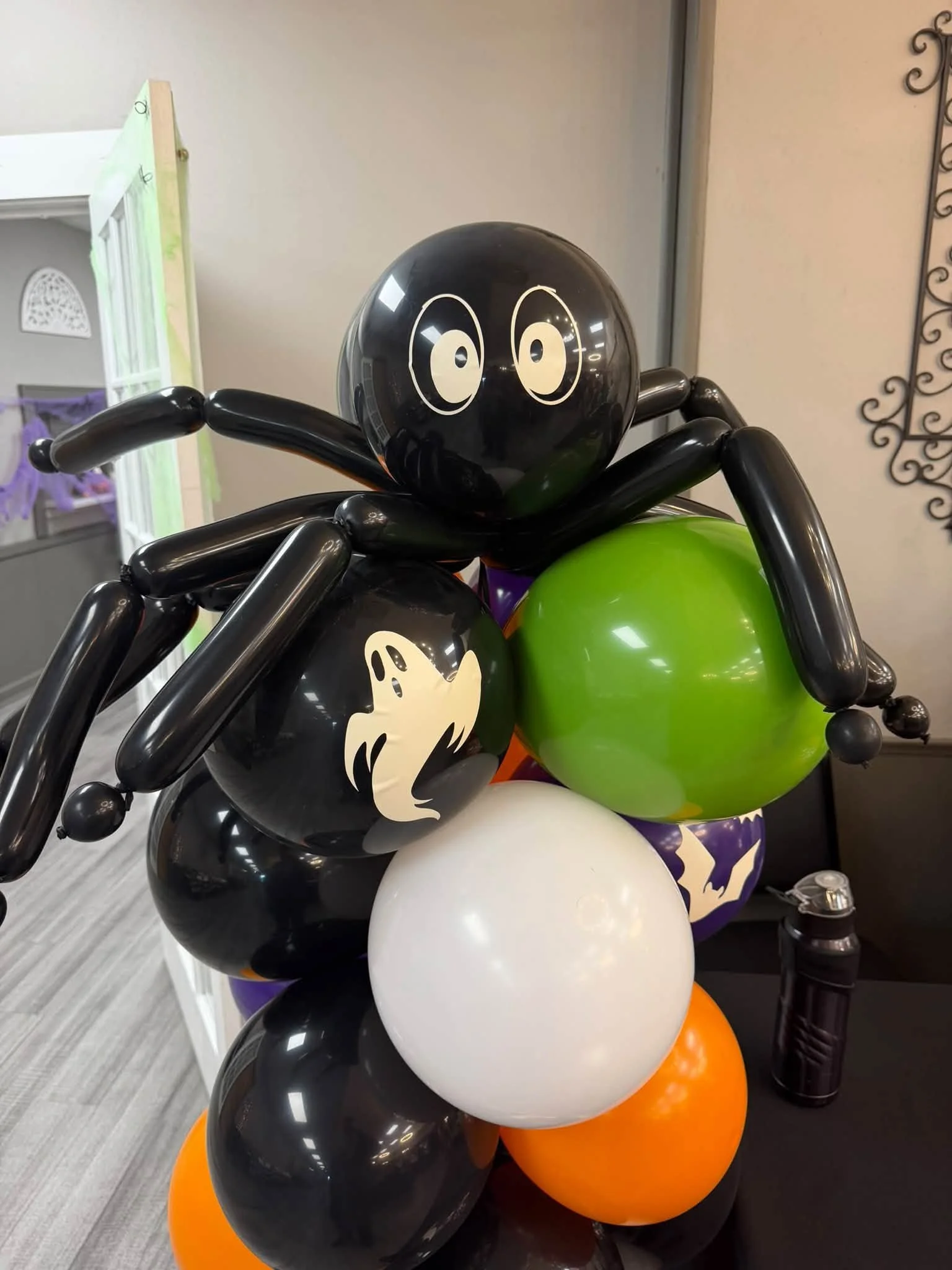 Halloween balloon sculpture featuring a black spider with cartoon eyes and a ghost, surrounded by black, green, white, purple, and orange balloons