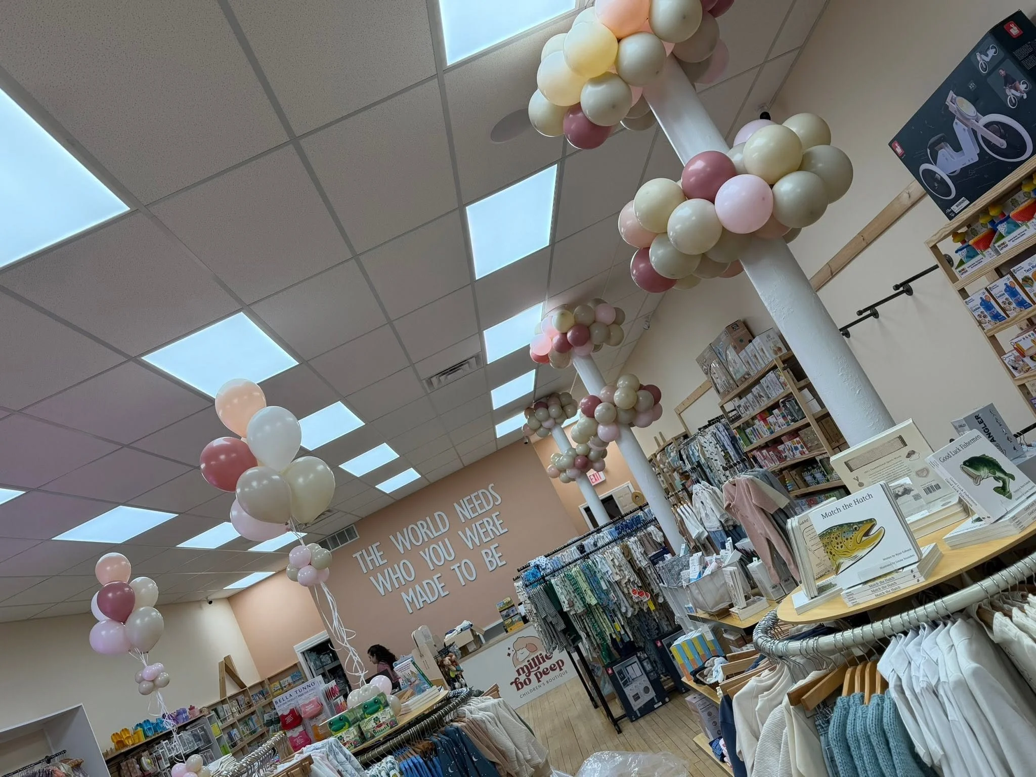 Interior of a children's boutique decorated with pastel balloon clusters, baby clothing racks, toys, books, and nursery products.