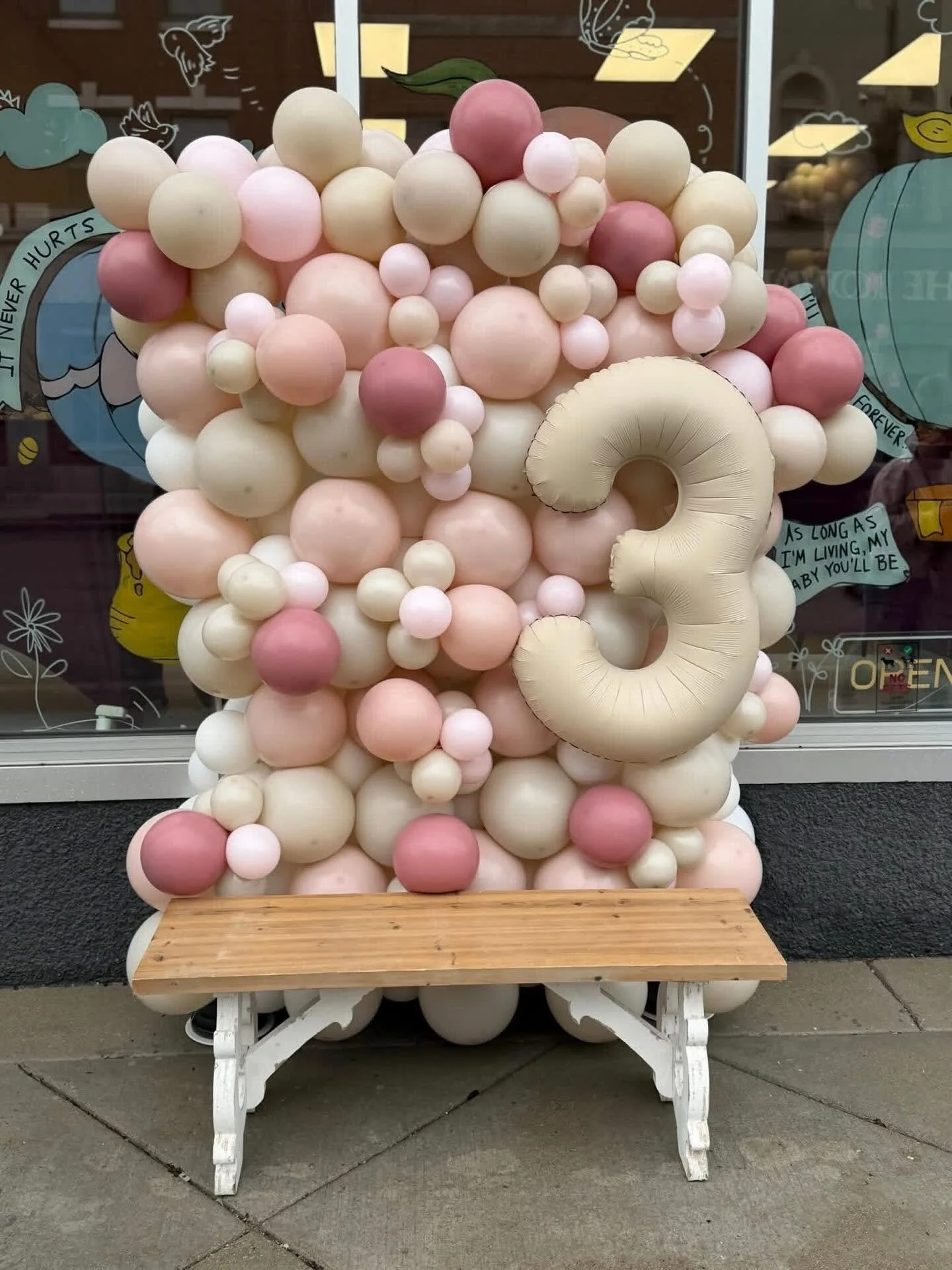 Pastel pink, cream, and white balloon arrangement with a large number 9 balloon displayed on a wooden bench outside a storefront.