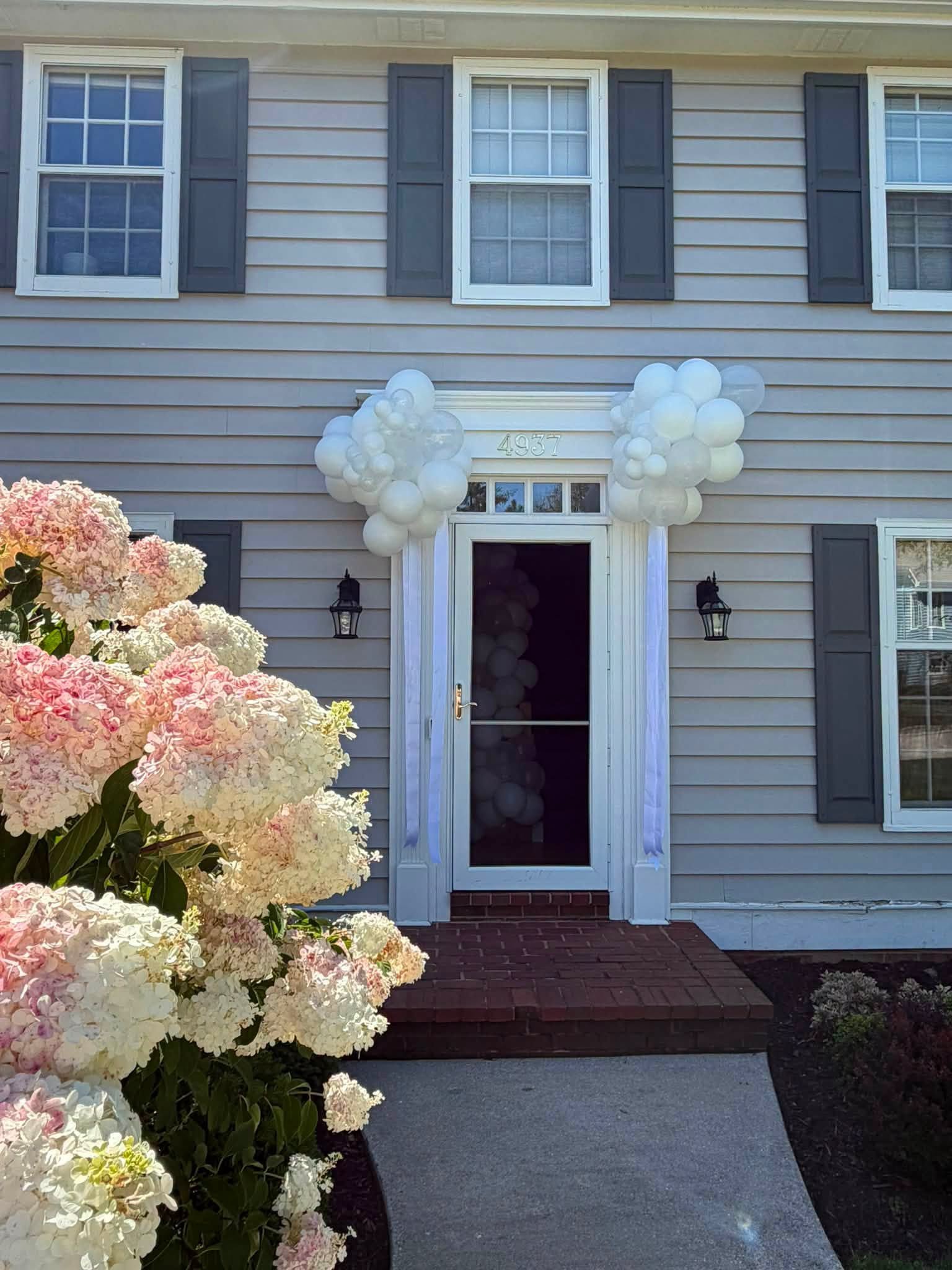 House entrance decorated with white balloon garlands, pink and white hydrangeas, and the address number 4937