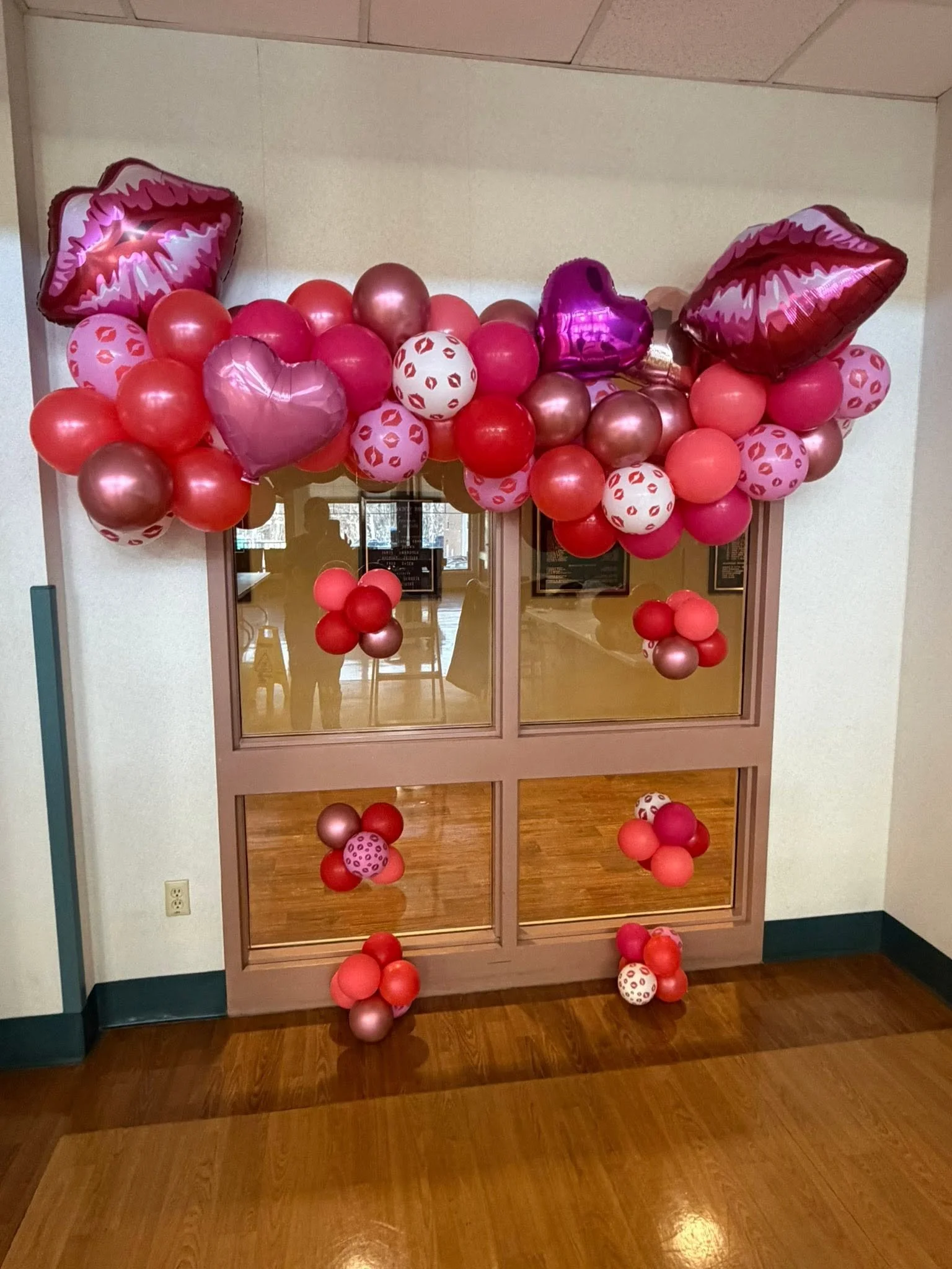 Pink and red balloon garland with heart, lips, and bear-shaped balloons decorating a glass doorway.