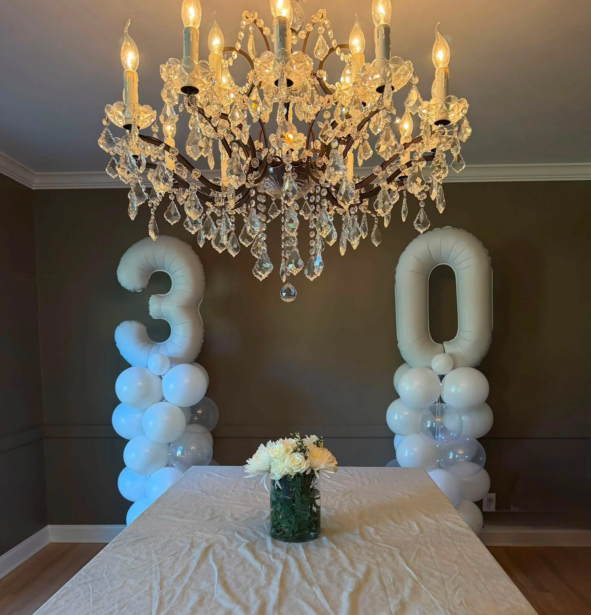 Elegant birthday table setting with a crystal chandelier, white balloon numbers 30, and a floral centerpiece.
