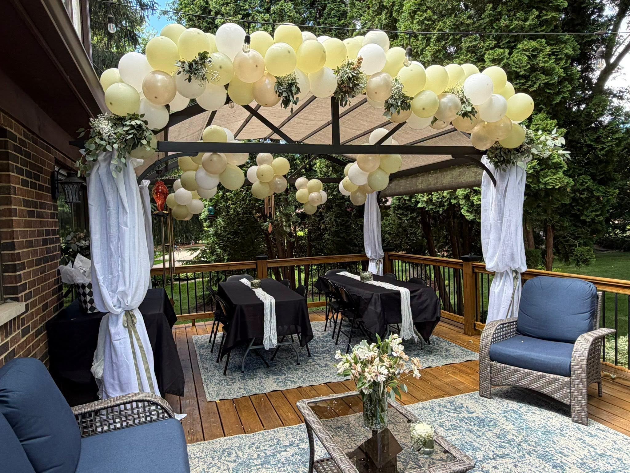 Outdoor wedding reception on a deck with a balloon arch, floral arrangements, draped pergola, and black-covered tables