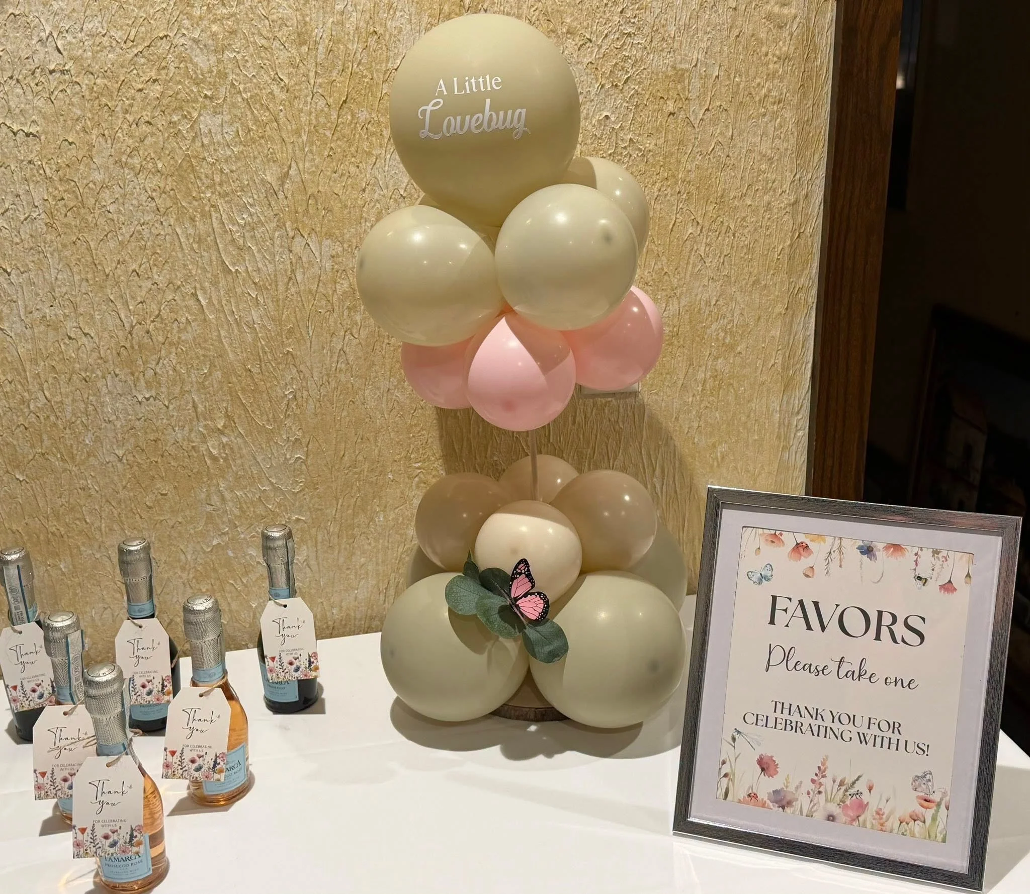 Wedding reception favor table with pastel balloon arrangement, mini wine bottles, and a framed “Favors Please take one” sign