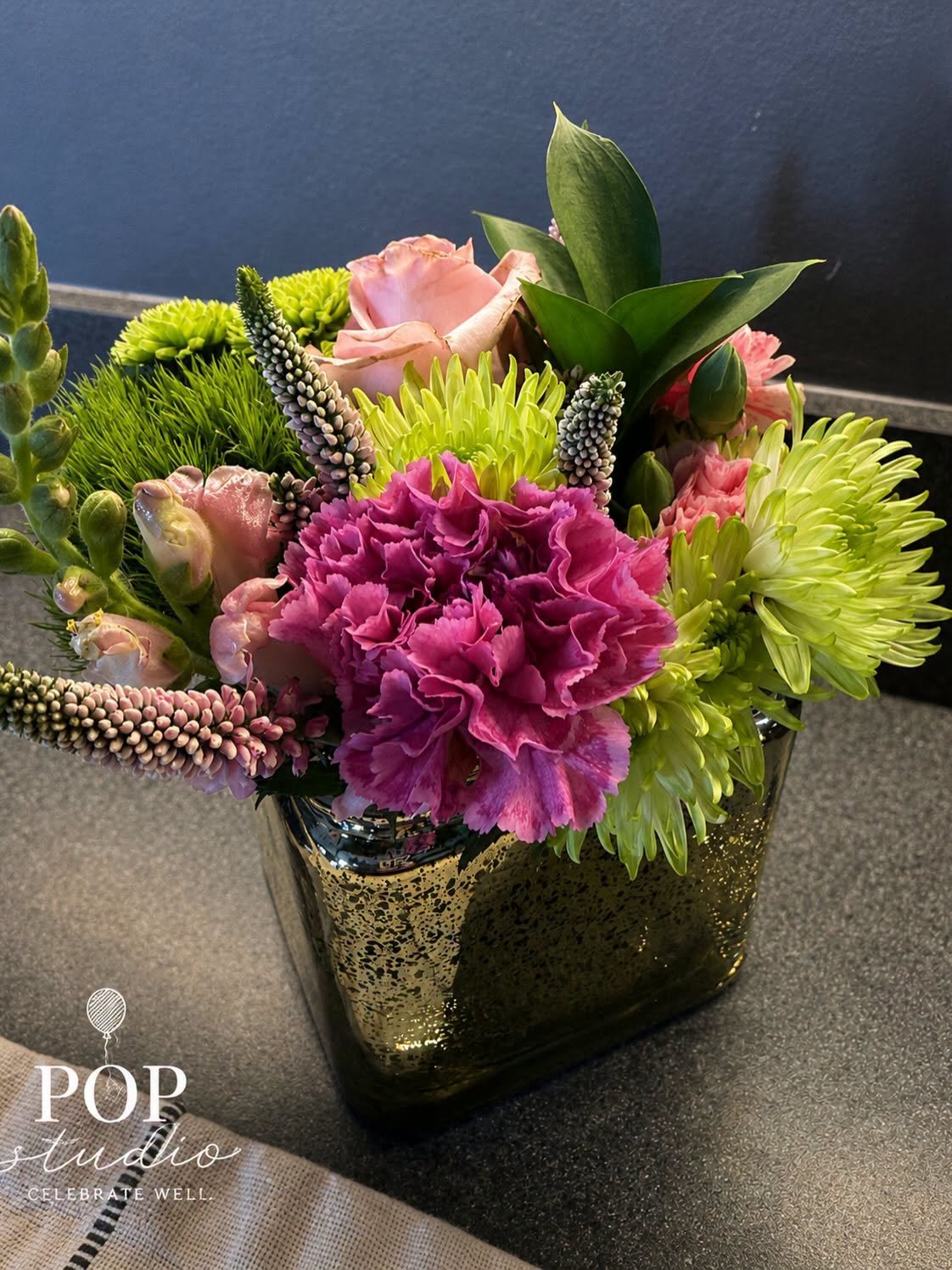 Colorful floral arrangement with pink roses, purple carnations, green chrysanthemums, and textured flowers in a gold-speckled vase