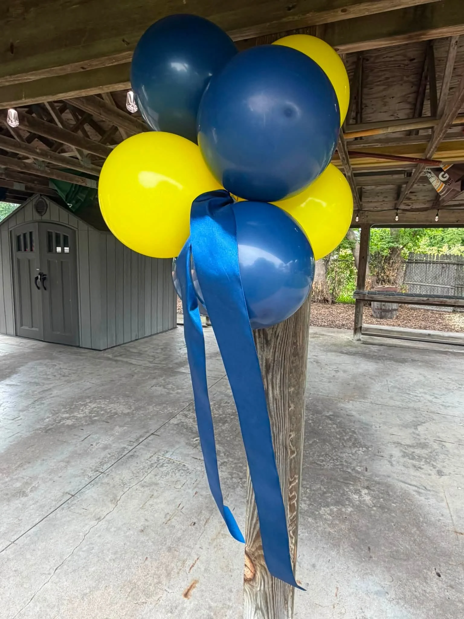 Large blue and yellow balloon sculpture with long blue ribbons under a covered outdoor pavilion