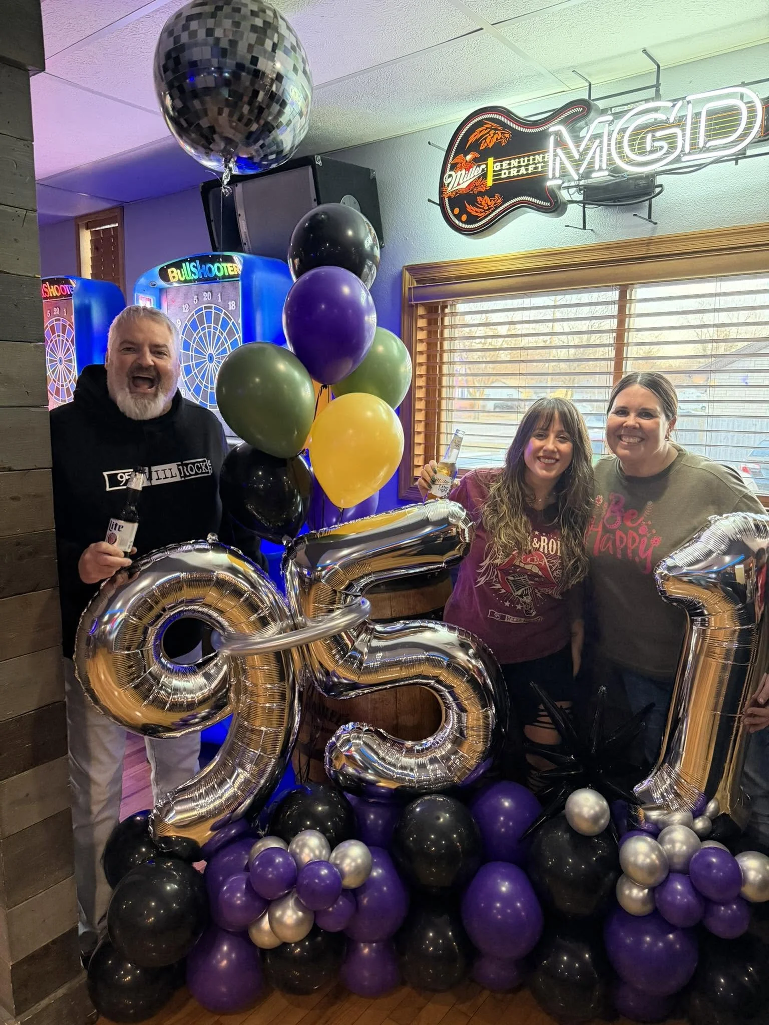Three people celebrating in a bar with colorful balloons, giant silver number balloons, a disco ball, and dartboards.