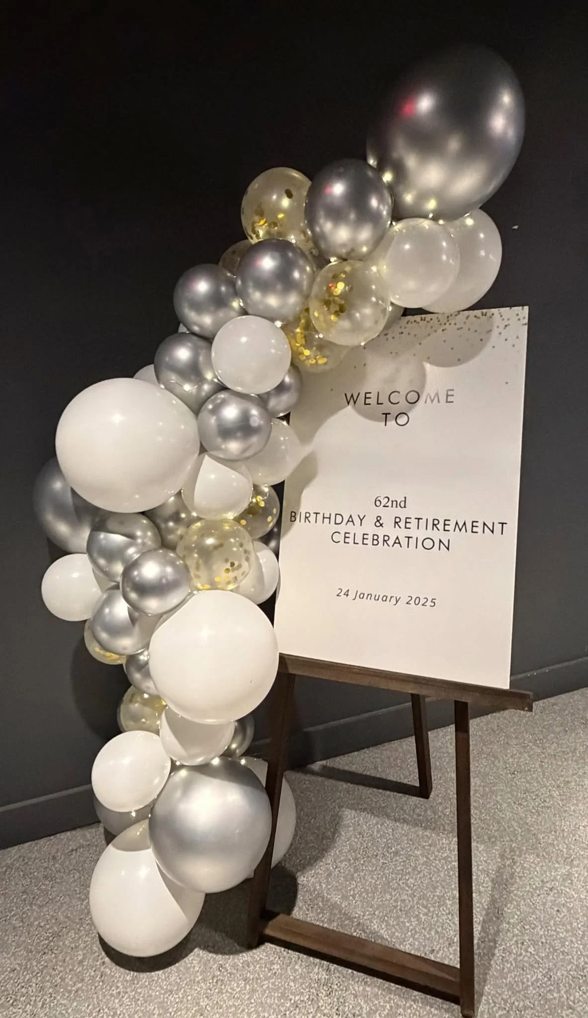 White, silver, and gold balloon garland beside a welcome sign for a 62nd birthday and retirement celebration.