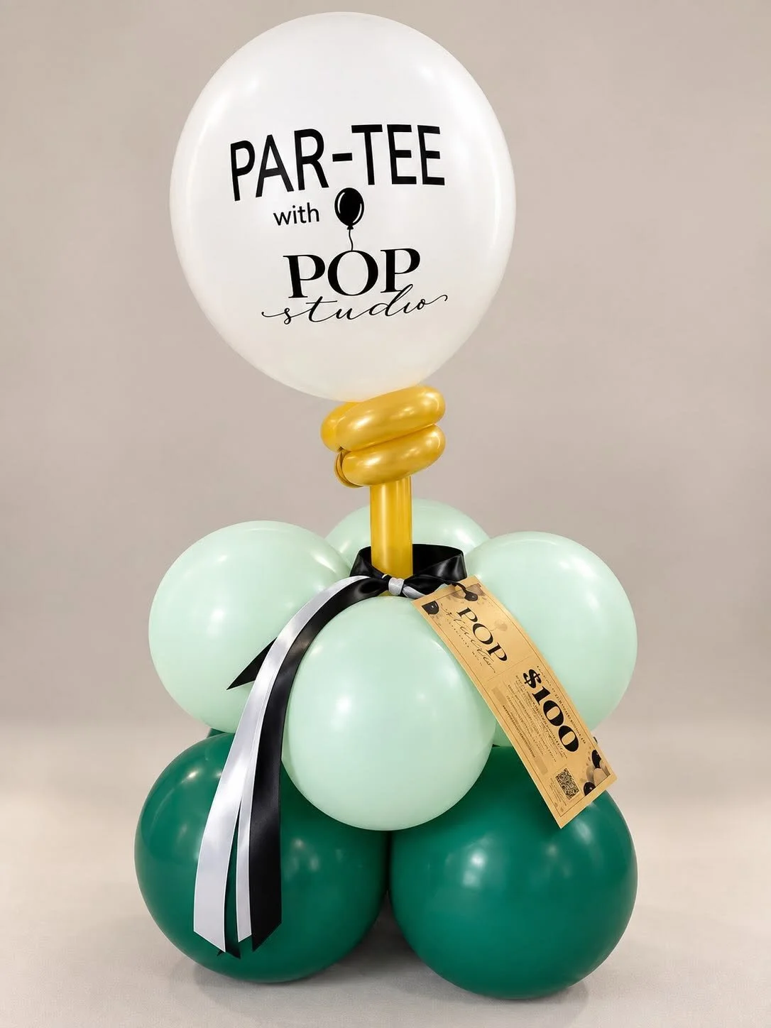 Decorative green and mint balloon arrangement with a gold balloon stem, black-and-white ribbon, and white balloon reading “PAR-TEE with POP studio.”