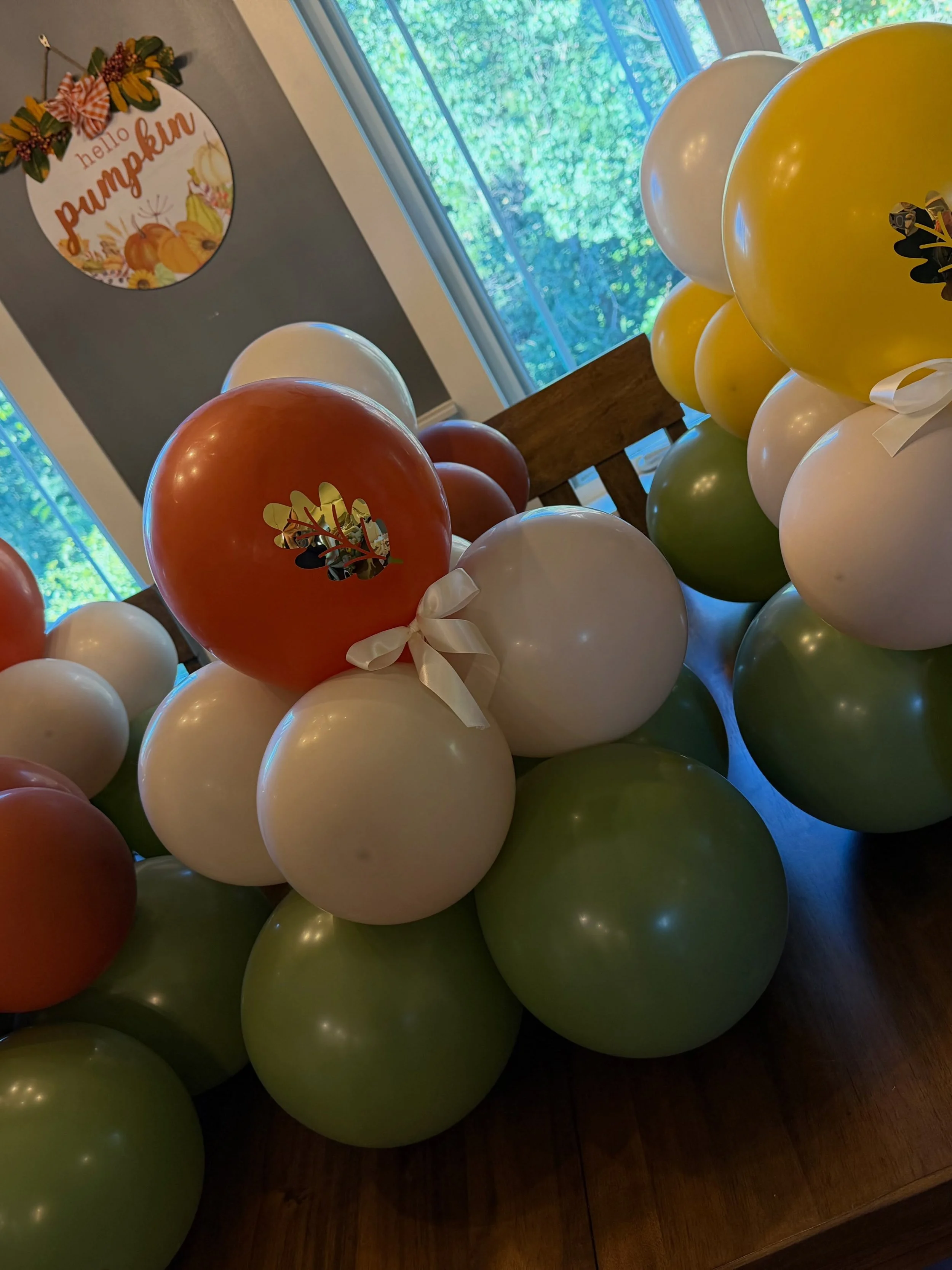 Autumn-themed balloon arrangement with orange, white, green, and yellow balloons beside a “hello pumpkin” floral wall sign.