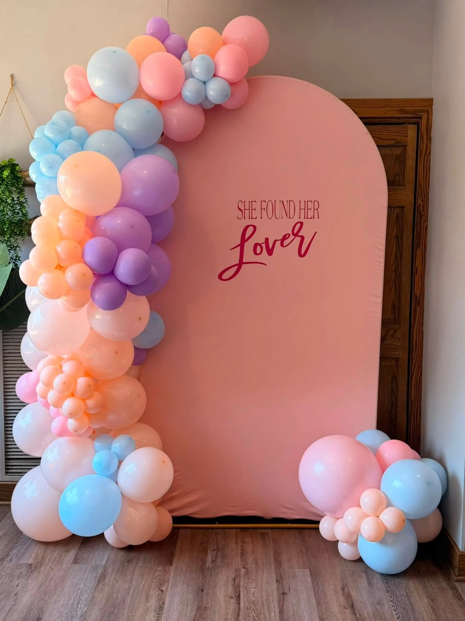 Pastel pink photo backdrop reading “She Found Her Lover,” framed by pink, lavender, peach, and blue balloon garlands.