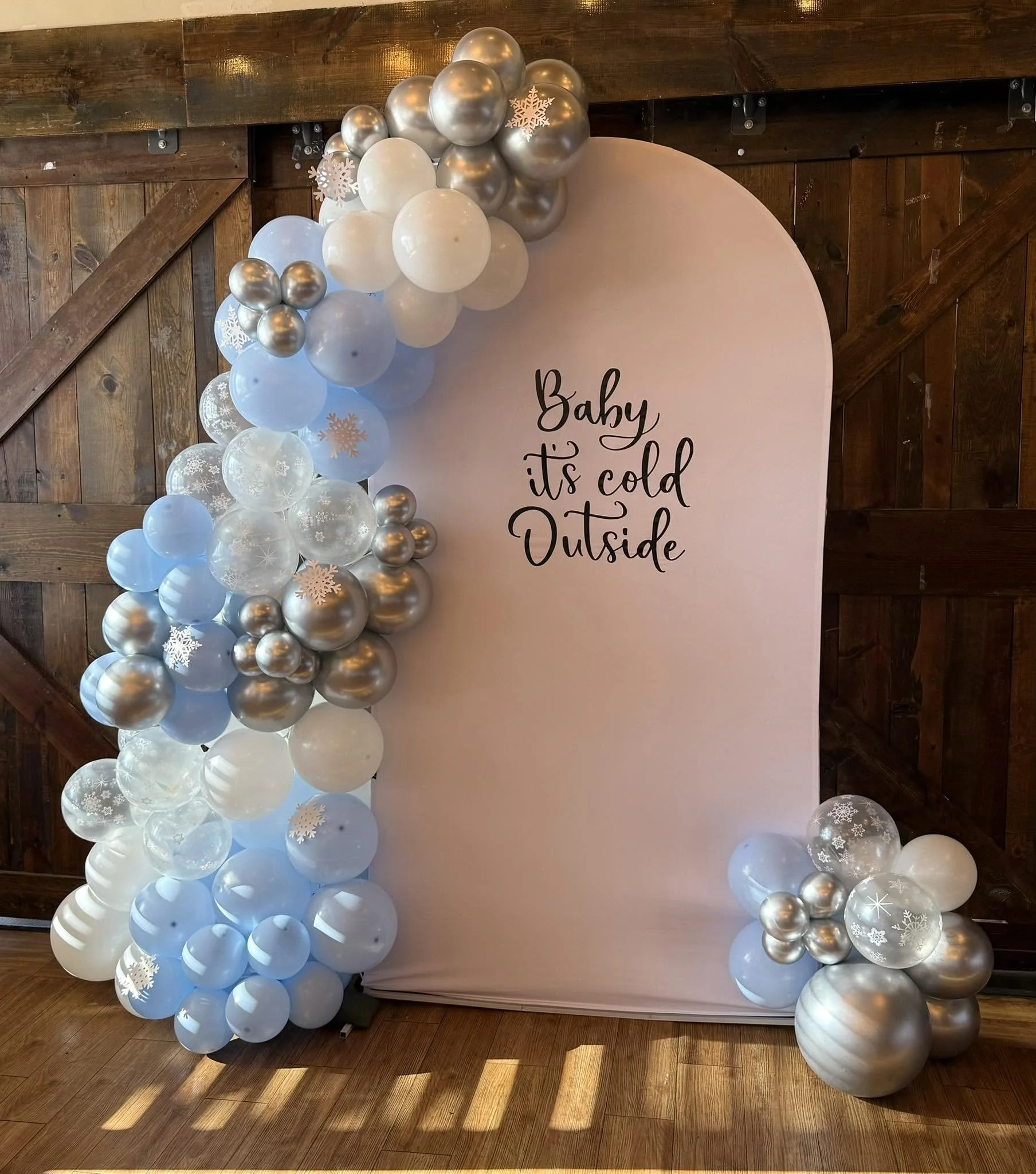 Winter-themed baby shower backdrop with blue, white, silver, and snowflake-decorated balloons surrounding a pink sign reading “Baby, it’s cold outside.”