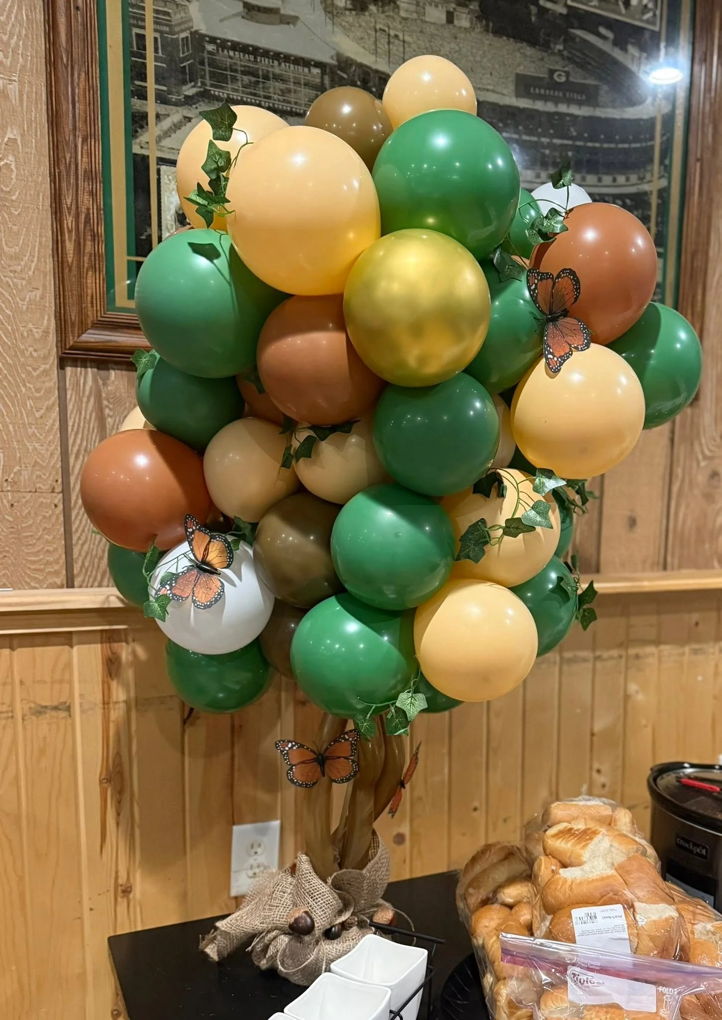 Colorful green, yellow, orange, and white balloon bouquet decorated with ivy leaves and butterfly ornaments indoors.
