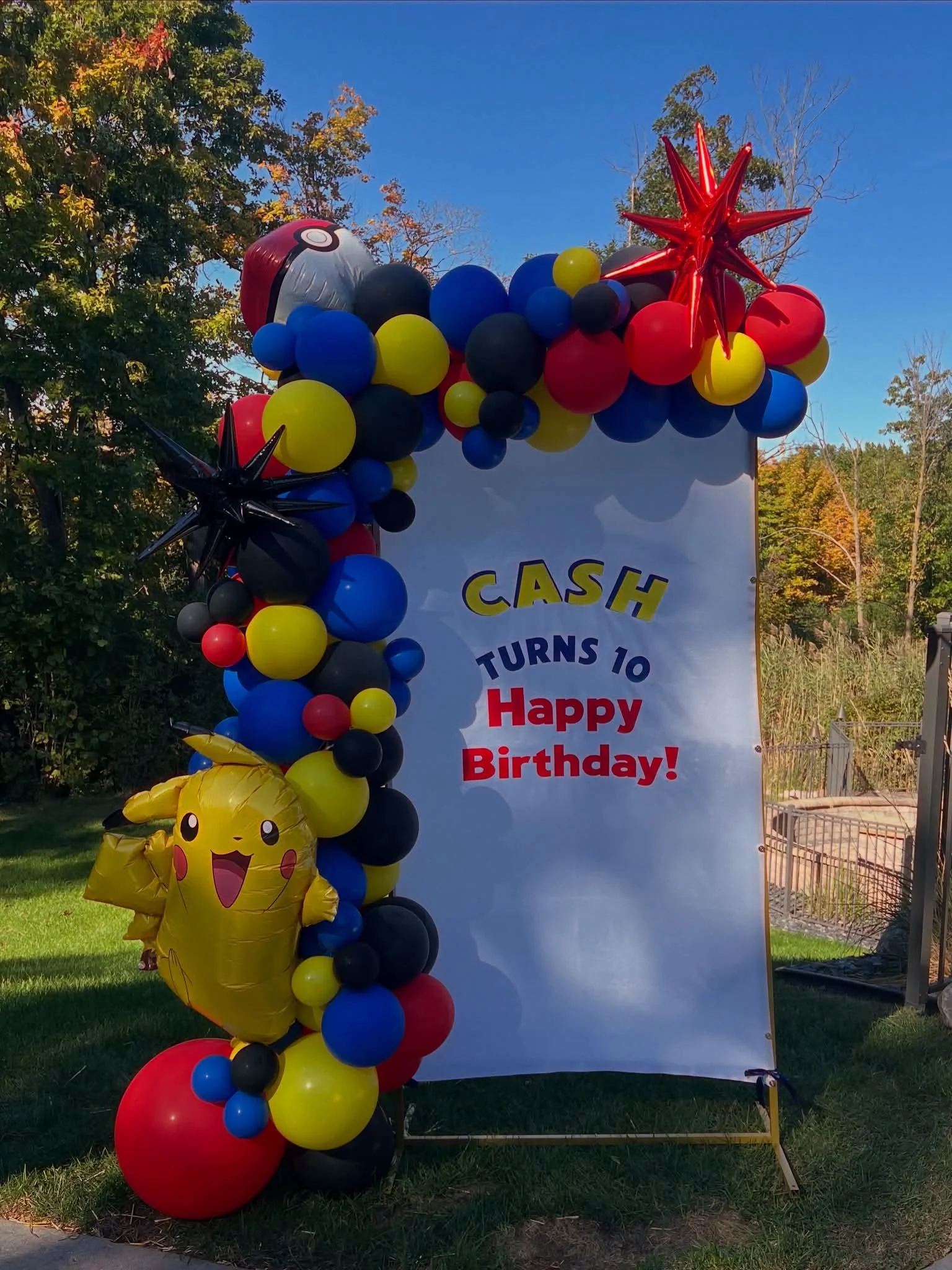 Colorful Pokémon-themed balloon arch with Pikachu and Poké Ball balloons framing a sign that reads “Cash turns 10. Happy Birthday!” outdoors.