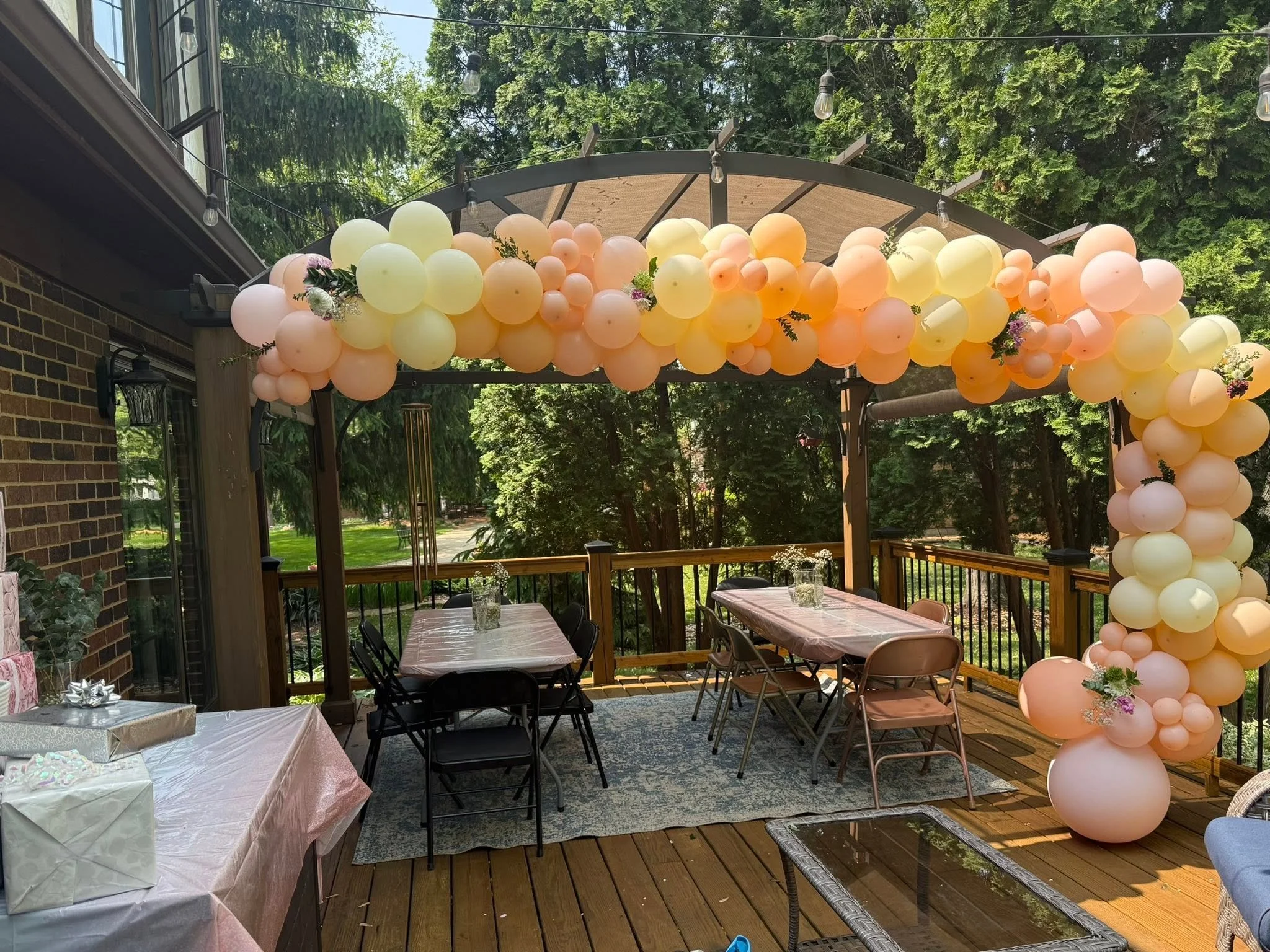 Outdoor party on a backyard deck with pastel peach, pink, and yellow balloon arch, decorated tables, string lights, and greenery.