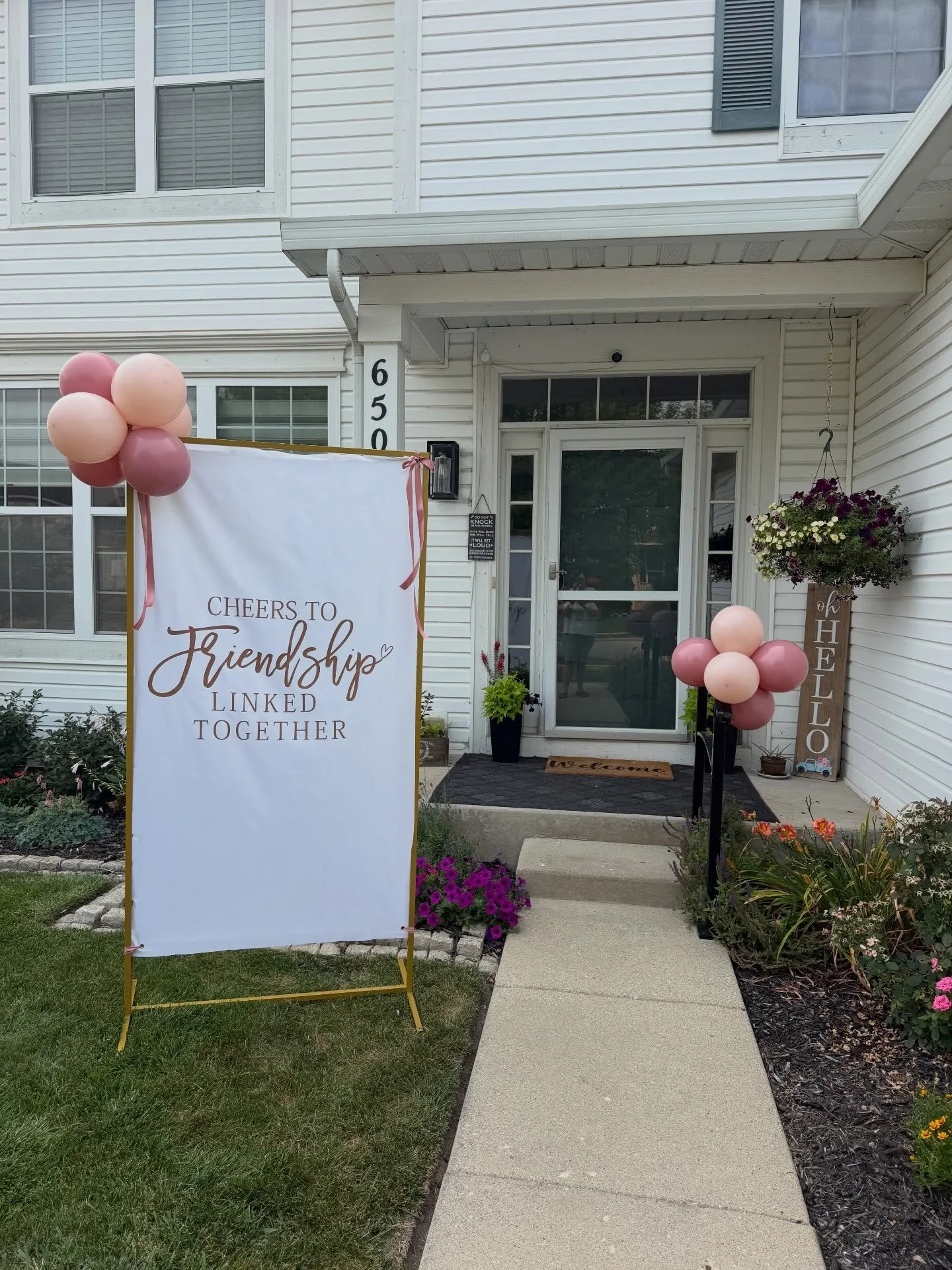 House entrance decorated with pink balloons, flowers, and a sign reading “Cheers to Friendship Linked Together.”