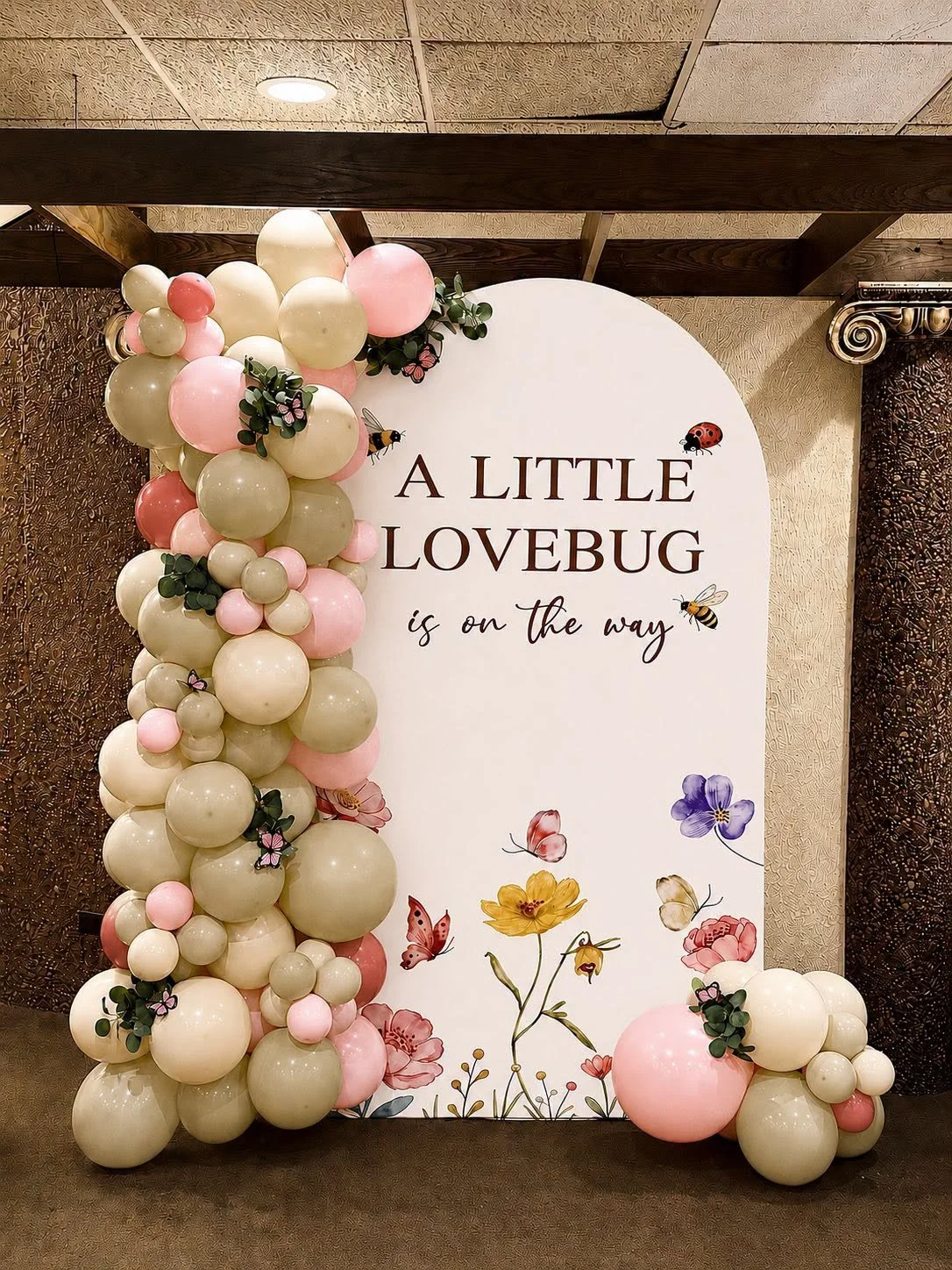 Baby shower welcome sign reading “A Little Lovebug is on the way,” framed by blush and cream balloon garlands, flowers, butterflies, bees, and a ladybug.