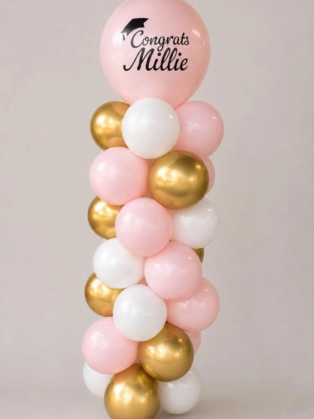 Pink, white, and gold balloon arrangement topped with a pink "Congrats Millie" graduation balloon.
