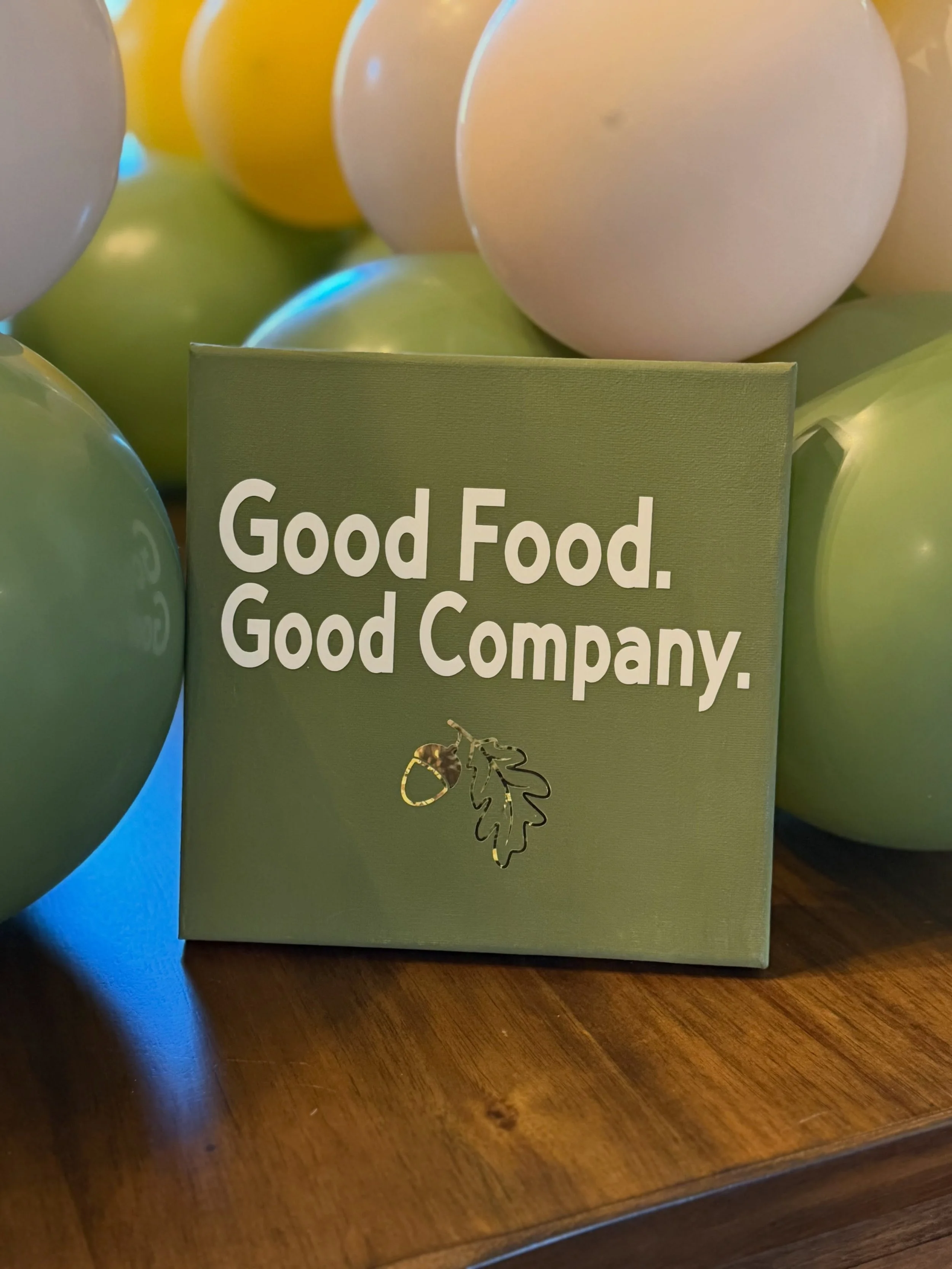 Green “Good Food. Good Company.” sign surrounded by colorful balloons on a wooden table.