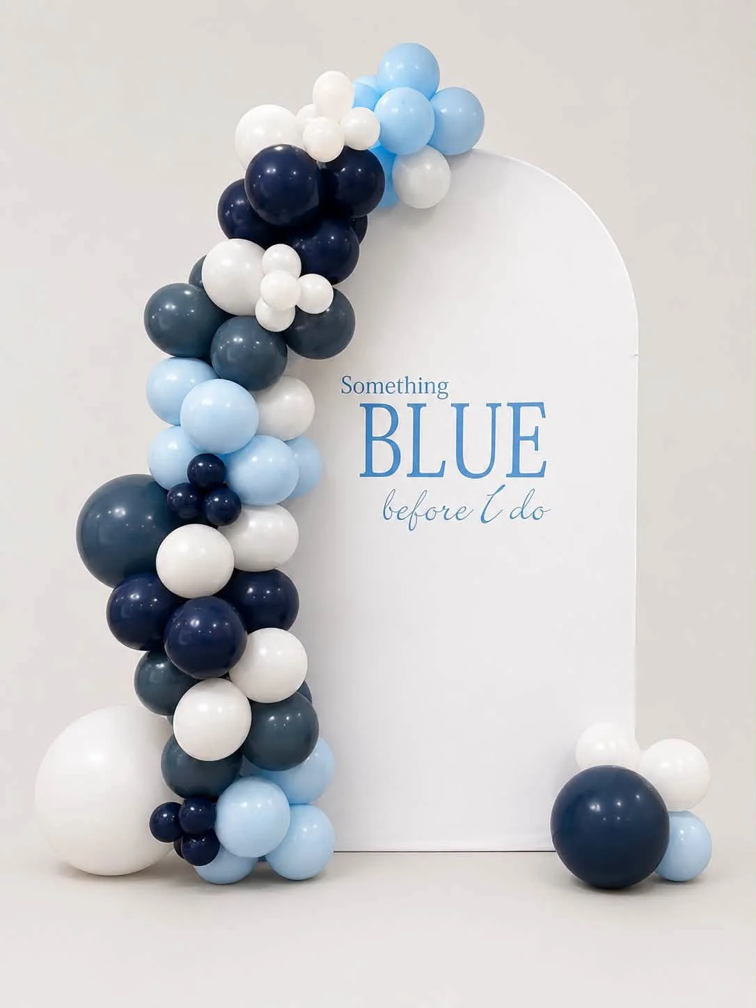 Blue, navy, white, and gray balloon arch framing a white sign reading “Something BLUE before I do,” with balloon clusters on the floor.
