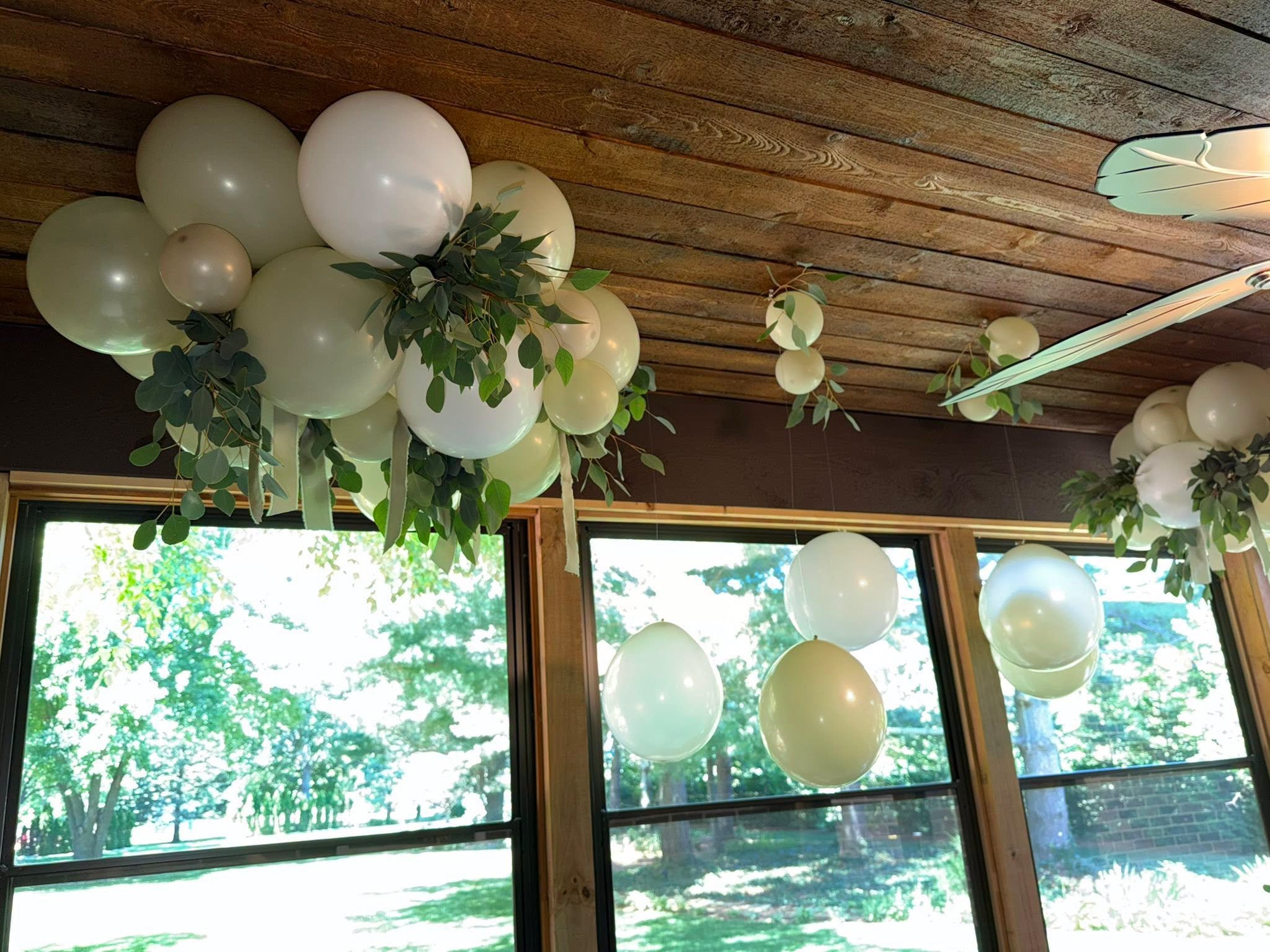 White balloon garlands with greenery and hanging paper decorations beneath a rustic wood ceiling.