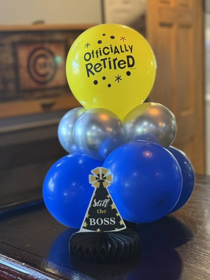 Retirement balloon arrangement with a yellow “Officially Retired” balloon, blue and silver balloons, and a “Still the Boss” party hat