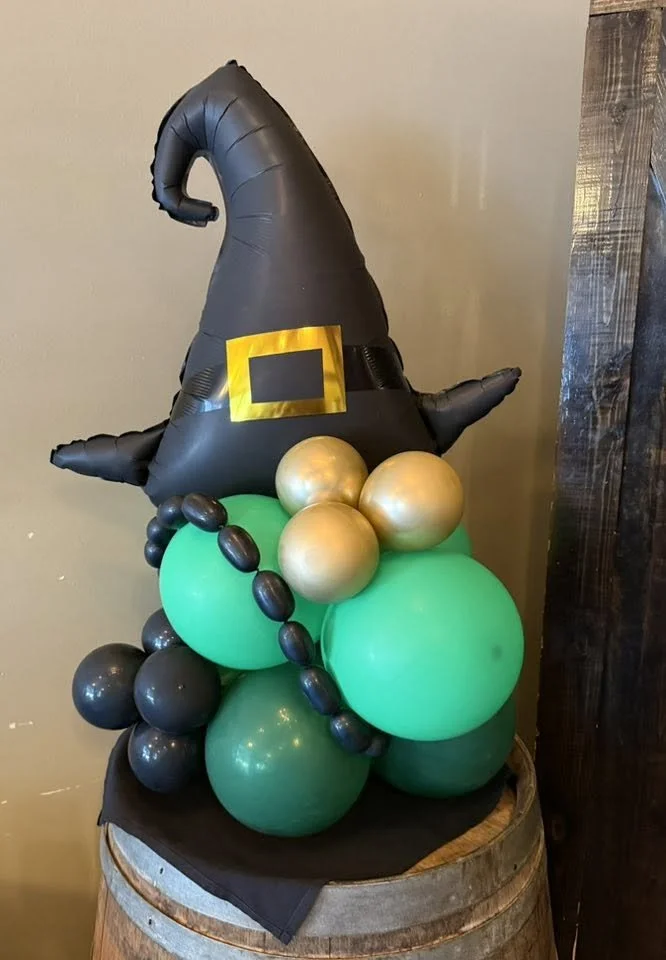 Balloon sculpture of a witch’s hat with gold buckle atop green, black, and gold balloons on a wooden barrel