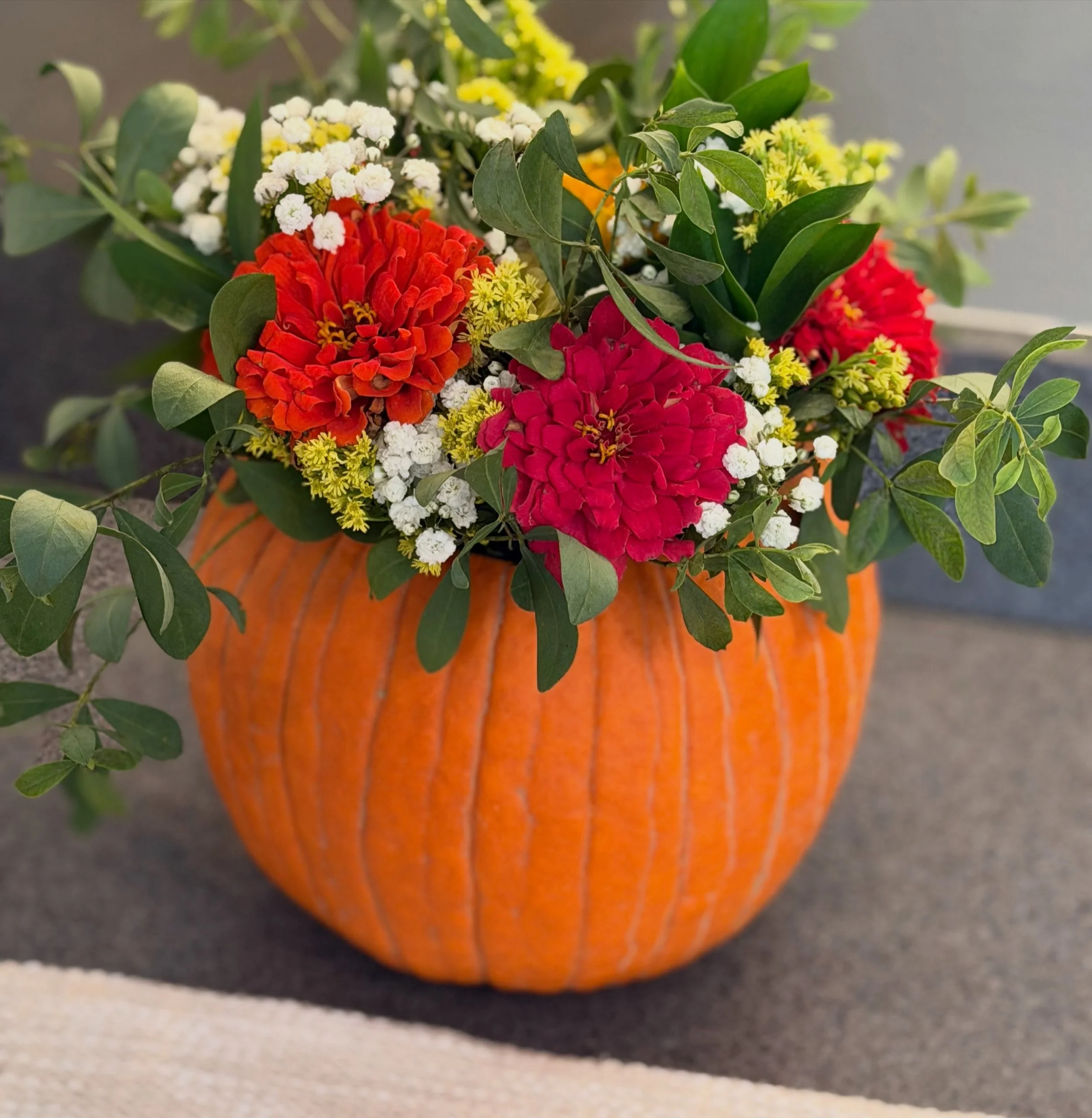 Colorful flower arrangement with red zinnias, white and yellow flowers, and greenery in an orange pumpkin vase