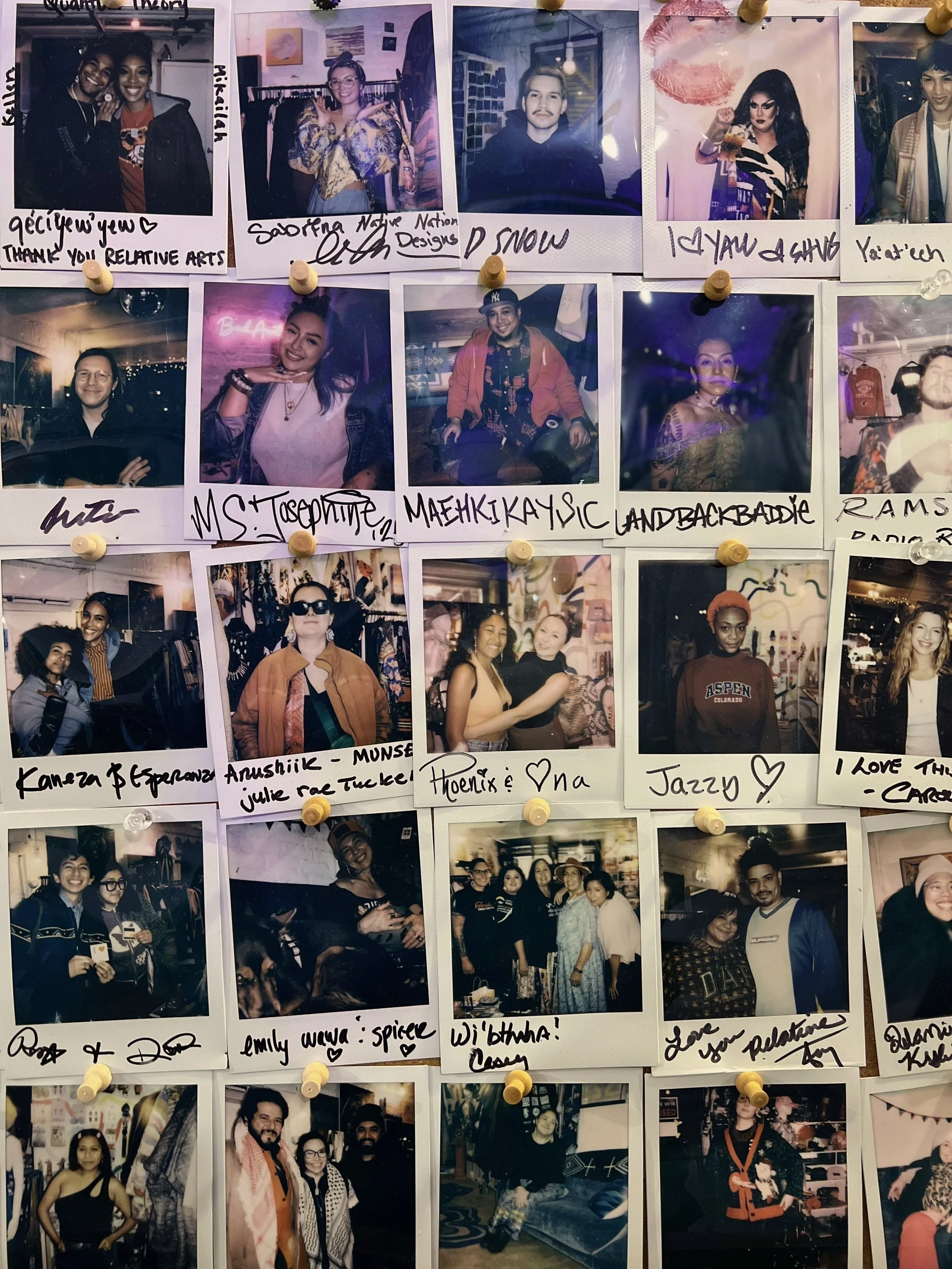 Wall covered with pinned Polaroid-style photographs of diverse people at social gatherings, each labeled with handwritten names and messages.