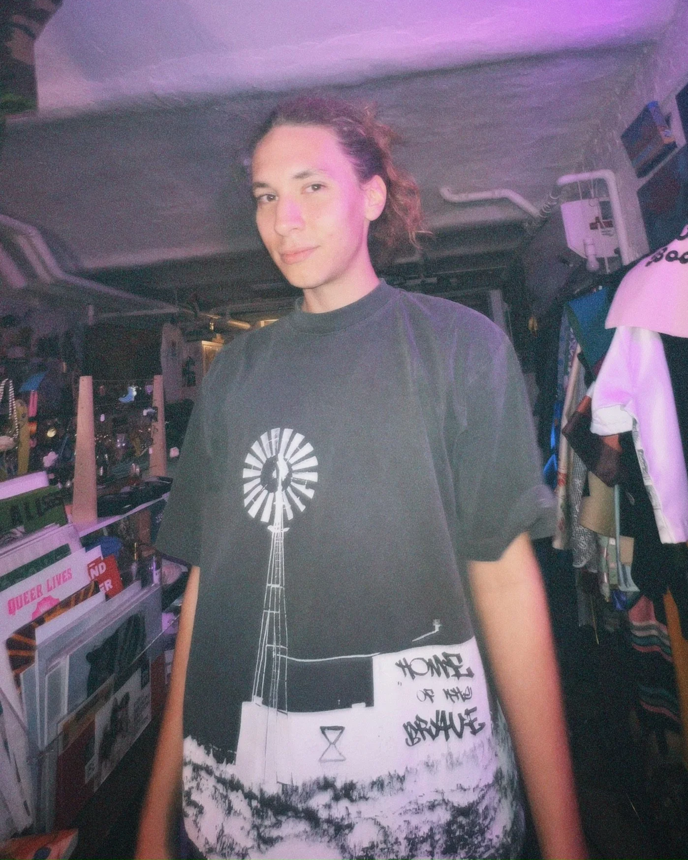 Person wearing a graphic black T-shirt standing in a dimly lit bedroom or closet surrounded by clothing and shelves.