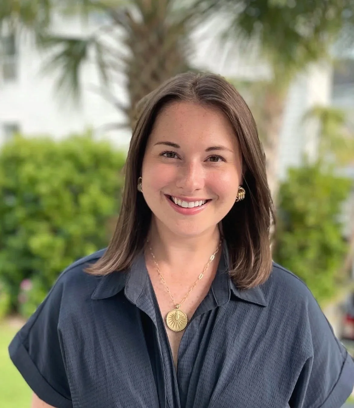 A smiling woman with shoulder-length brown hair, wearing a dark blue shirt, gold jewelry, and a gold necklace, standing outdoors with green trees and plants in the background.