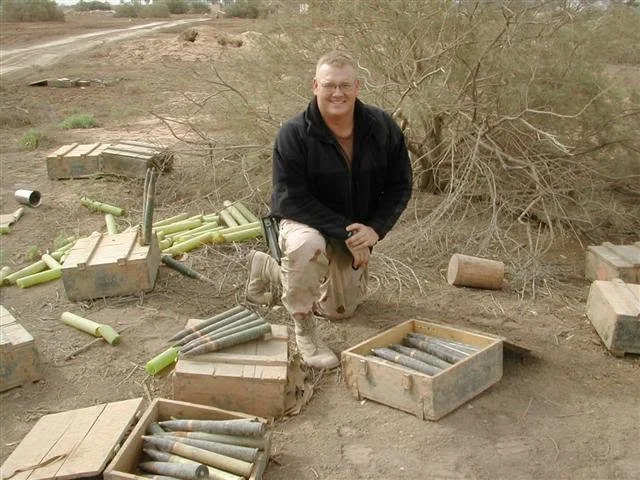 Man crouching outdoors with unexploded artillery shells and discharged shells around him.