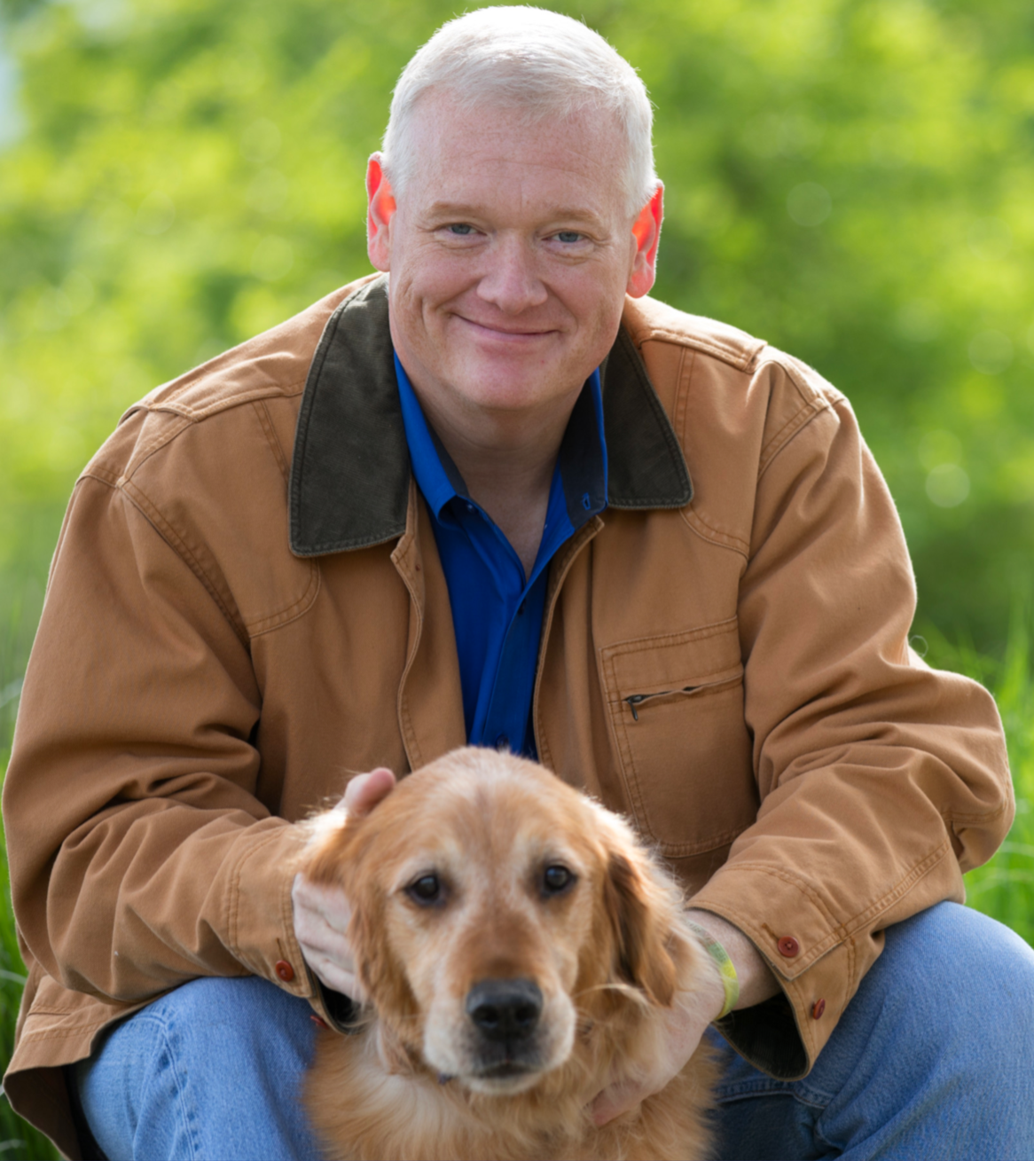 A man with short gray hair and a blue shirt sitting outdoors with a golden retriever puppy in front of him, both looking at the camera.