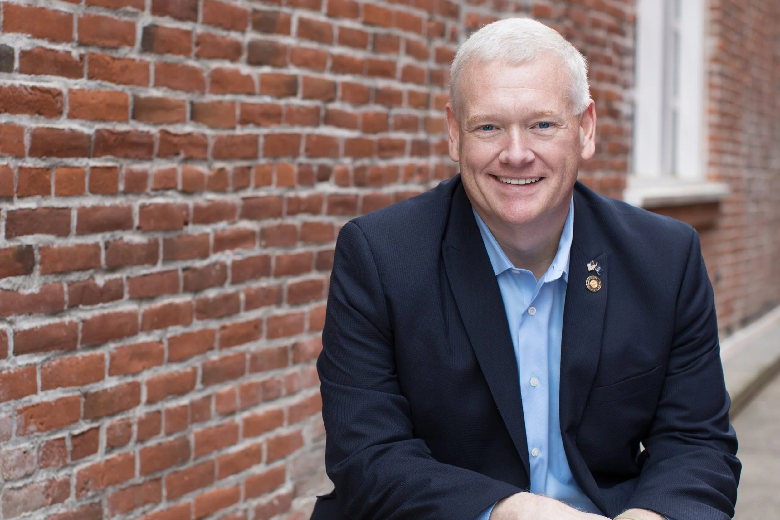 A smiling man with white hair and blue eyes wearing a dark suit and light blue shirt, sitting in front of a red brick wall.