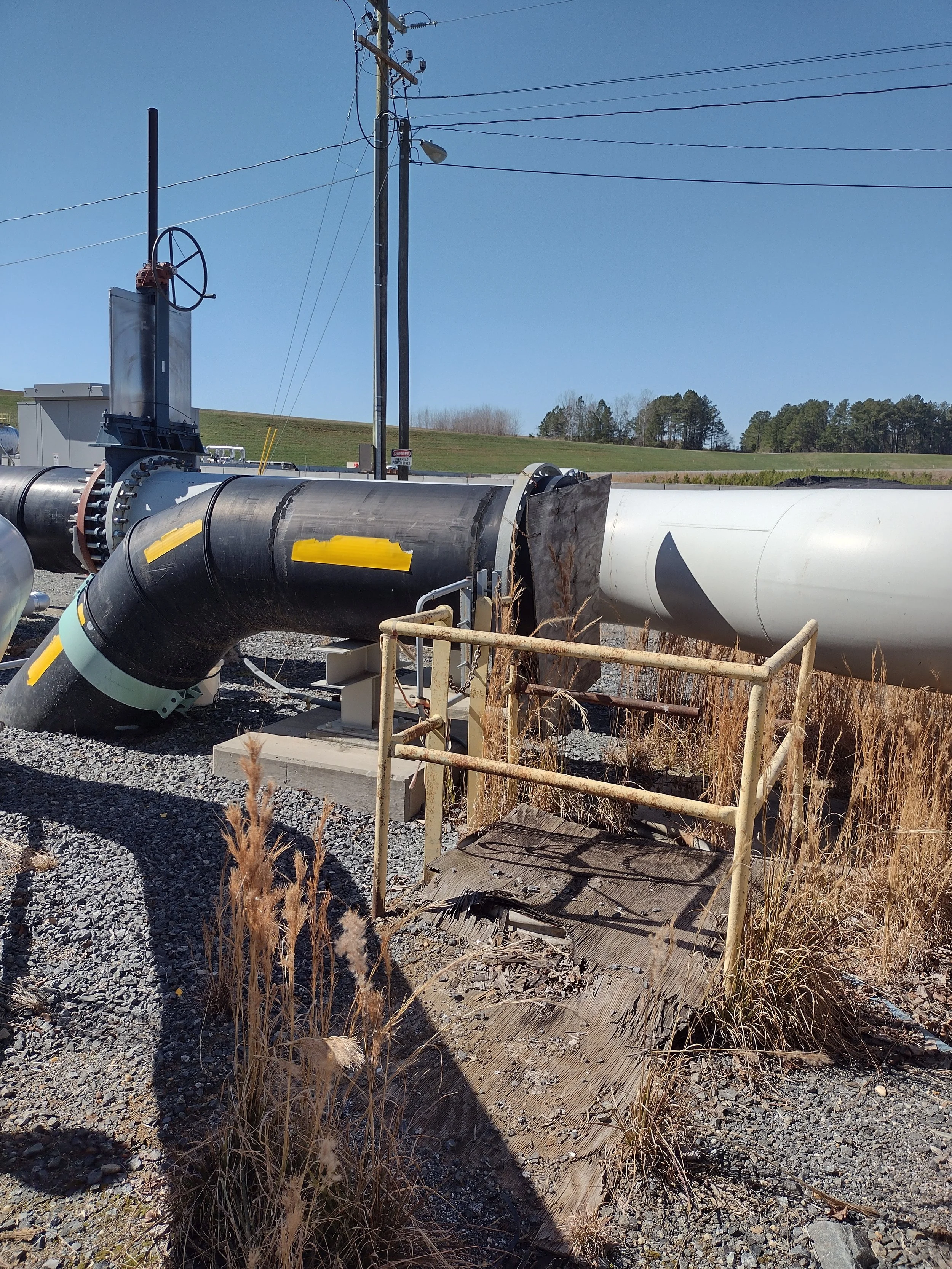 Large pipeline with black and white sections lying on gravel ground, with a wooden board and dry grass in the foreground, utility poles and wires, and green hills with trees in the background under a clear blue sky.